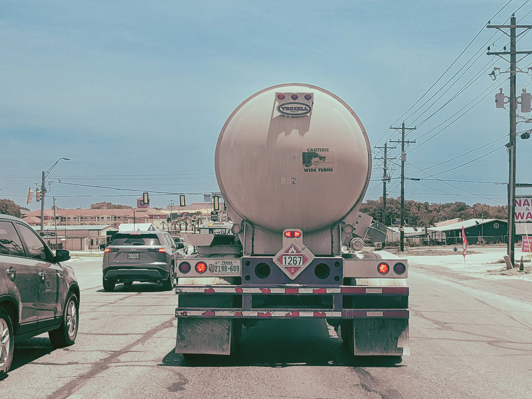 View of a large tanker truck in traffic surrounded by cars, utility poles, and buildings in the background on a cloudy day.