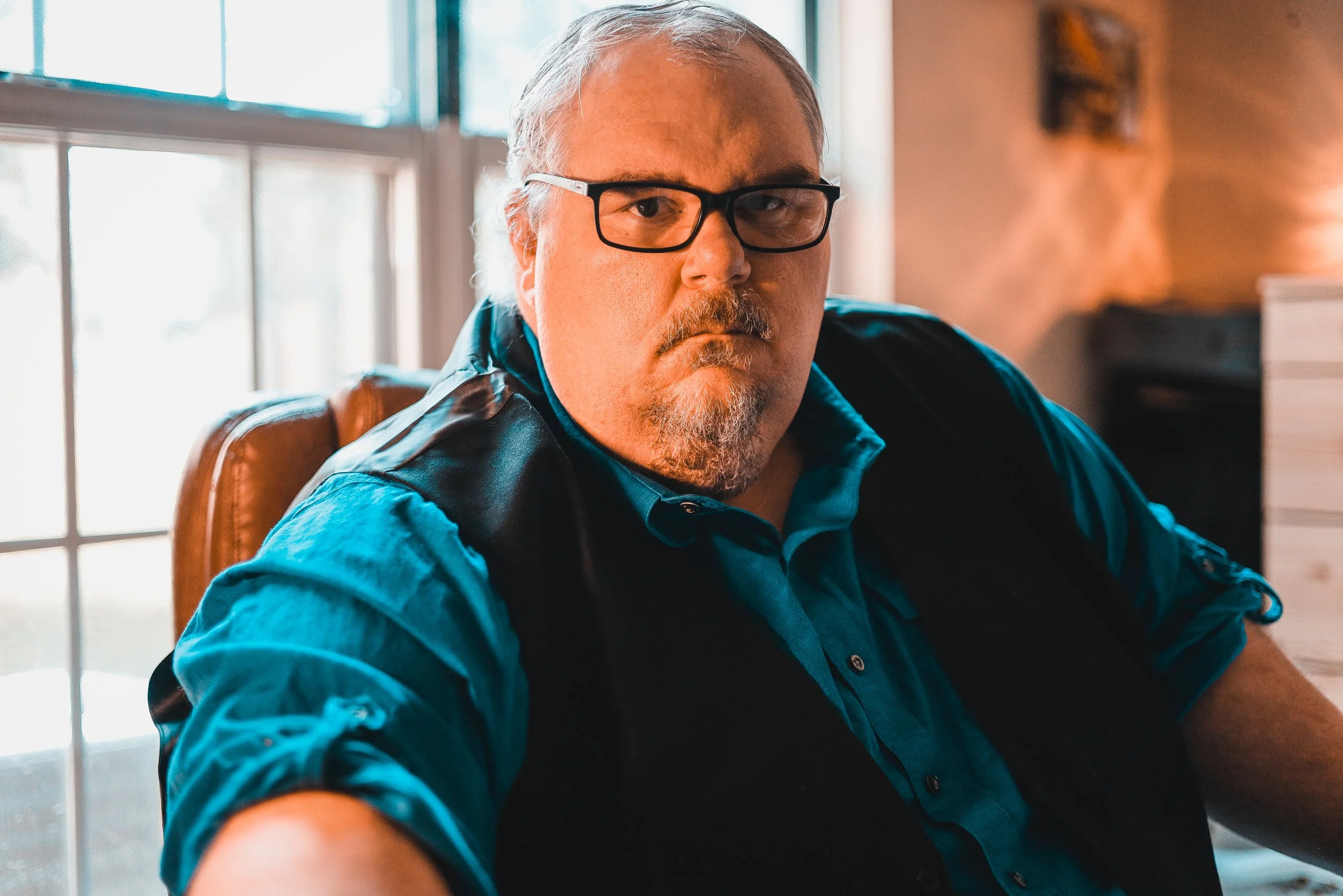 A middle-aged man with gray hair and a beard, wearing glasses, sitting indoors near a window with white frames and frosted glass, looking directly at the camera. He's dressed in a blue shirt and a black vest, with natural light illuminating his face.