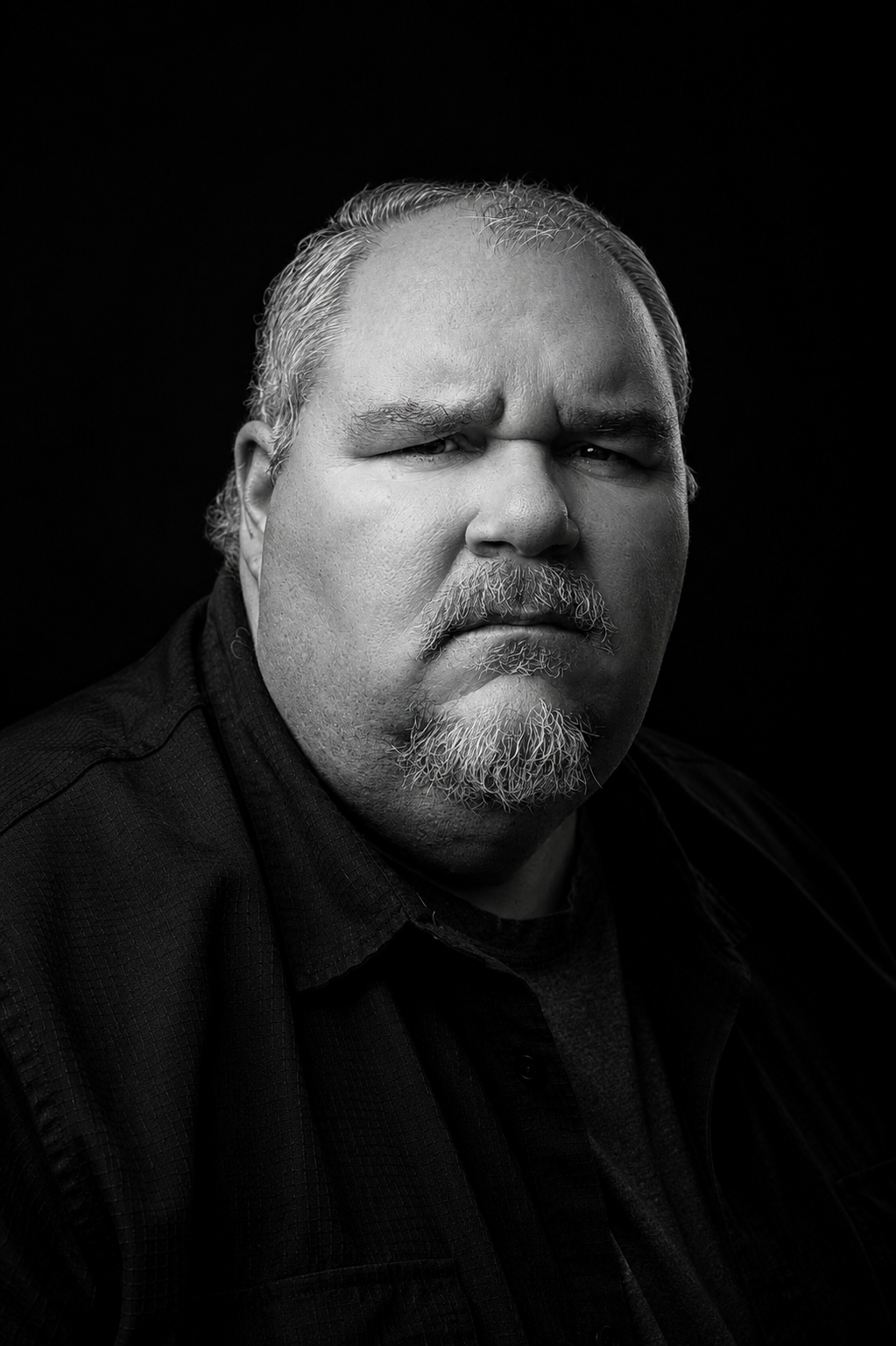 Black and white close-up portrait of a middle-aged man with a serious expression, light facial hair, and slicked-back hair against a dark background.