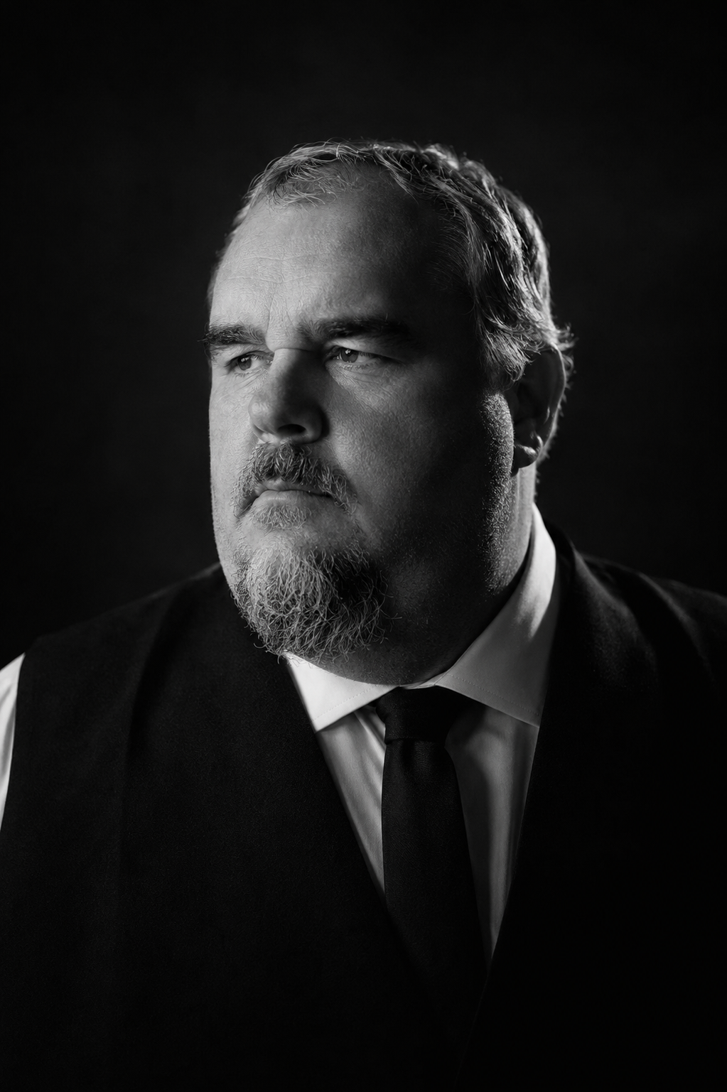 Black and white portrait of a man with a beard and mustache, wearing a suit vest, white shirt, and black tie, looking to the side against a dark background.