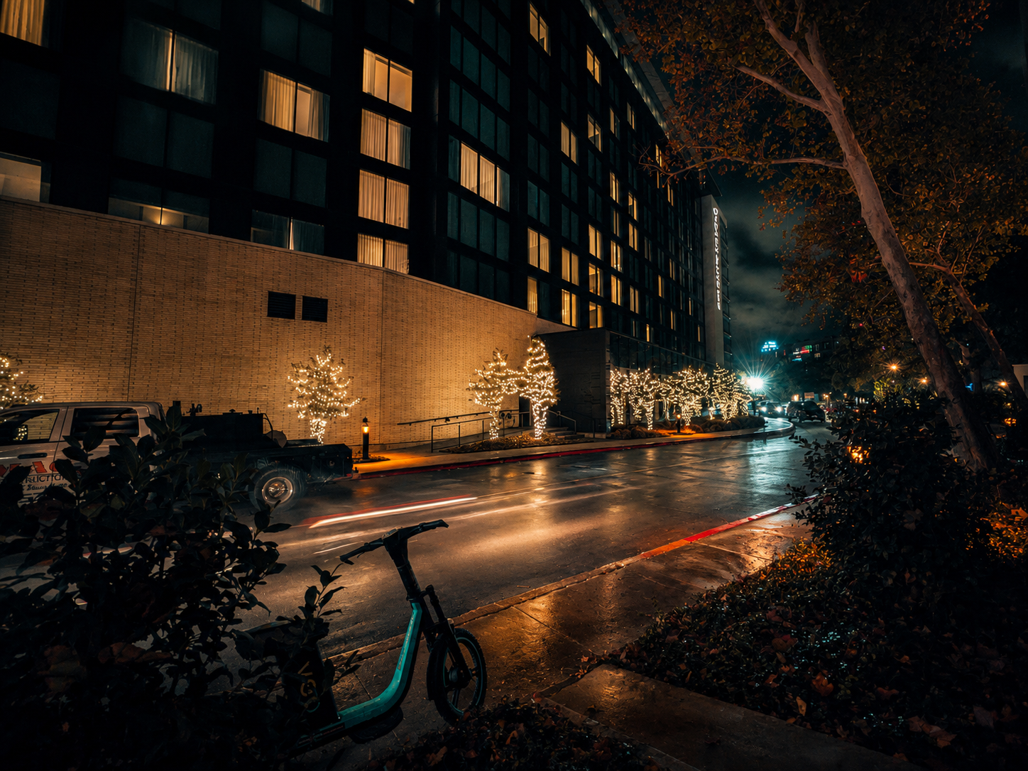 Nighttime city street with lit Christmas trees along sidewalk, wet road reflecting lights, parked cars, a bicycle in foreground, and a modern building with illuminated windows in the background.