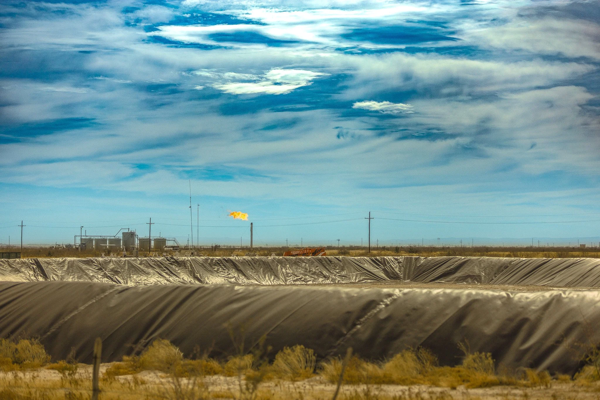An industrial landscape featuring a long, curved black barrier in the foreground, an open field with sparse vegetation, utility poles, and a building with a faint smoke or steam plume, all under a partly cloudy sky.