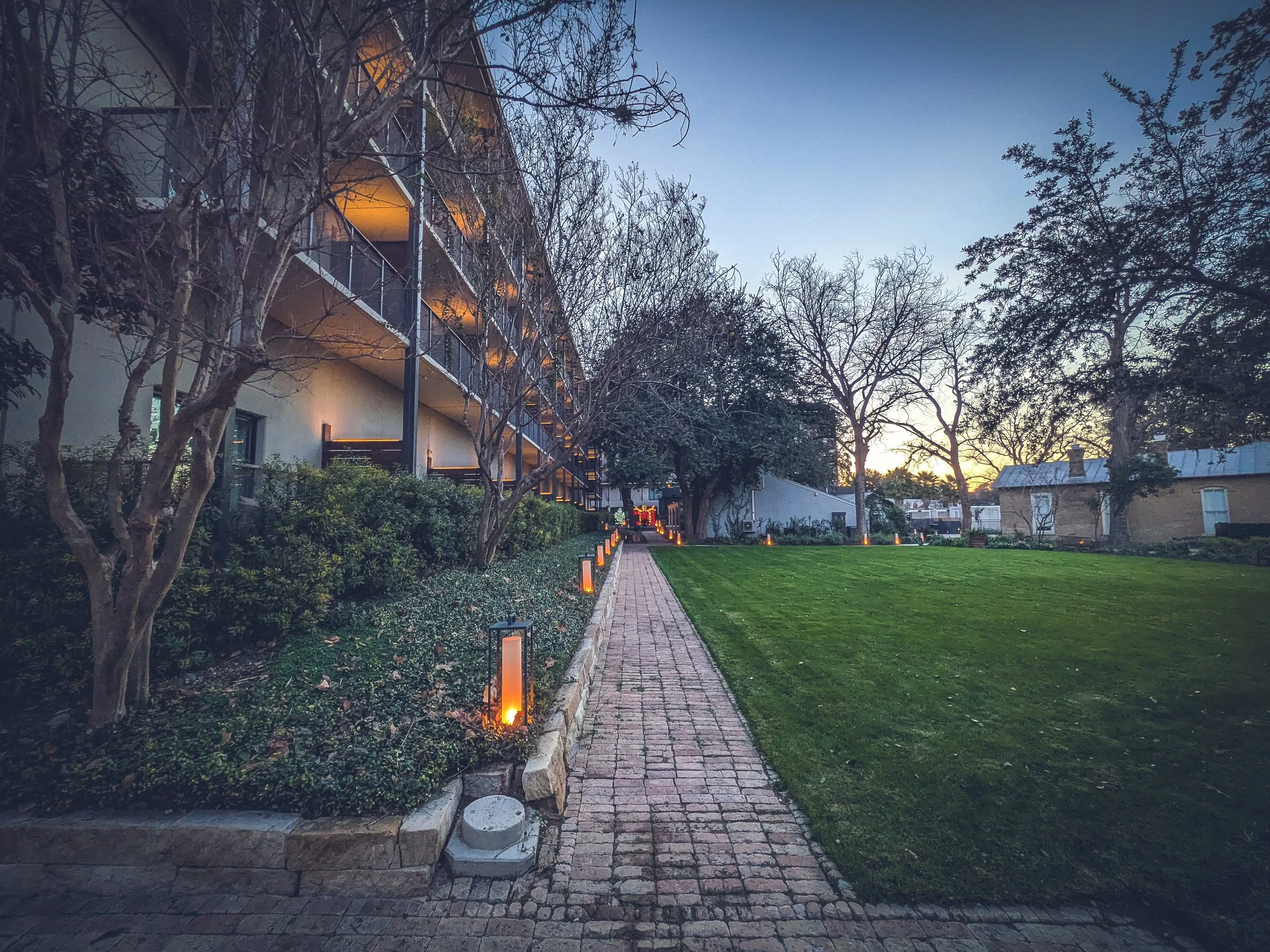 A landscaped brick pathway lined with lantern-style lights leads past a modern, multi-story hotel building with balconies, set against a manicured lawn and trees at dusk. Warm interior lighting contrasts with the cool tones of the early evening sky.