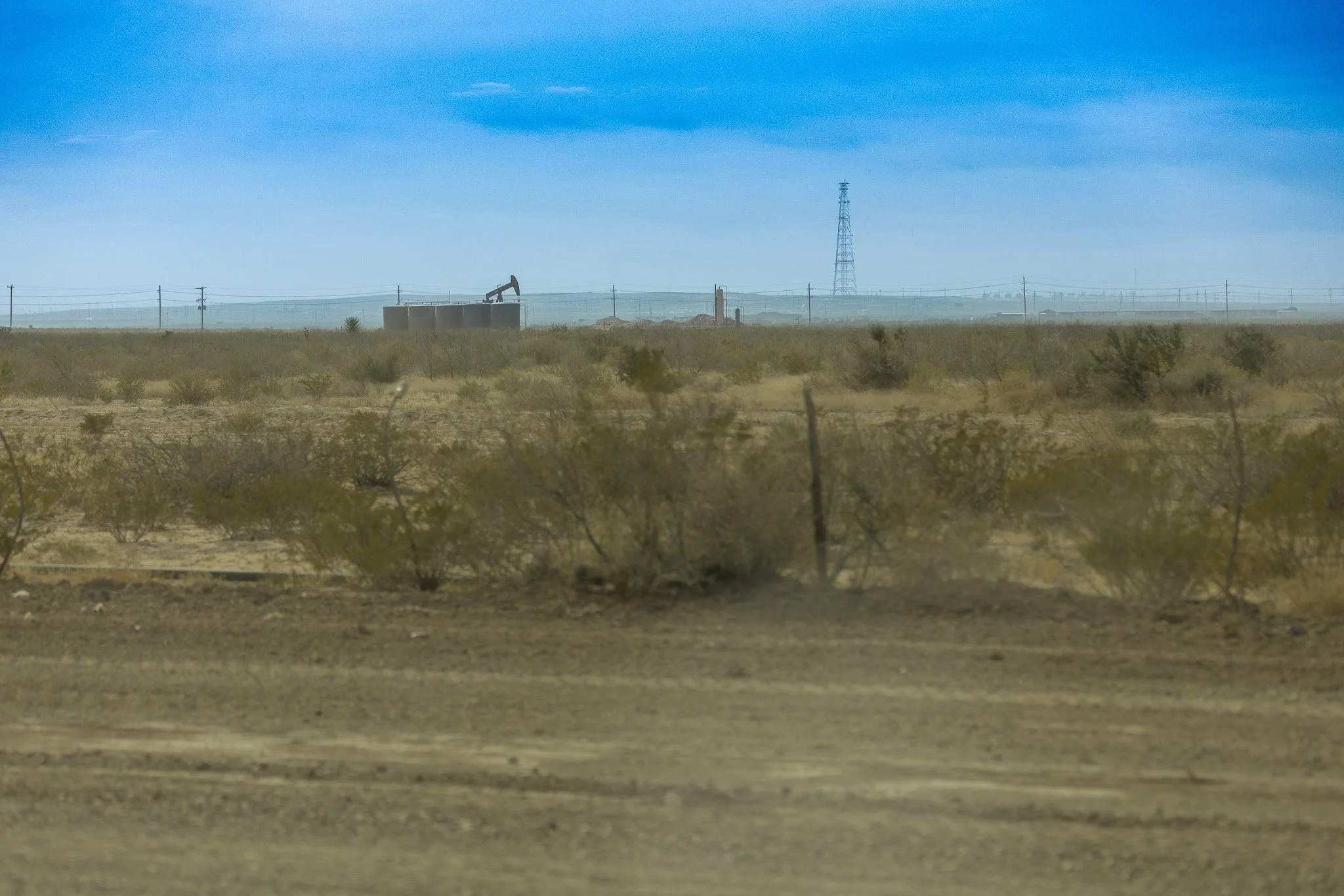Desert landscape with an oil pumpjack, power lines, and a transmission tower under a blue sky with some clouds.