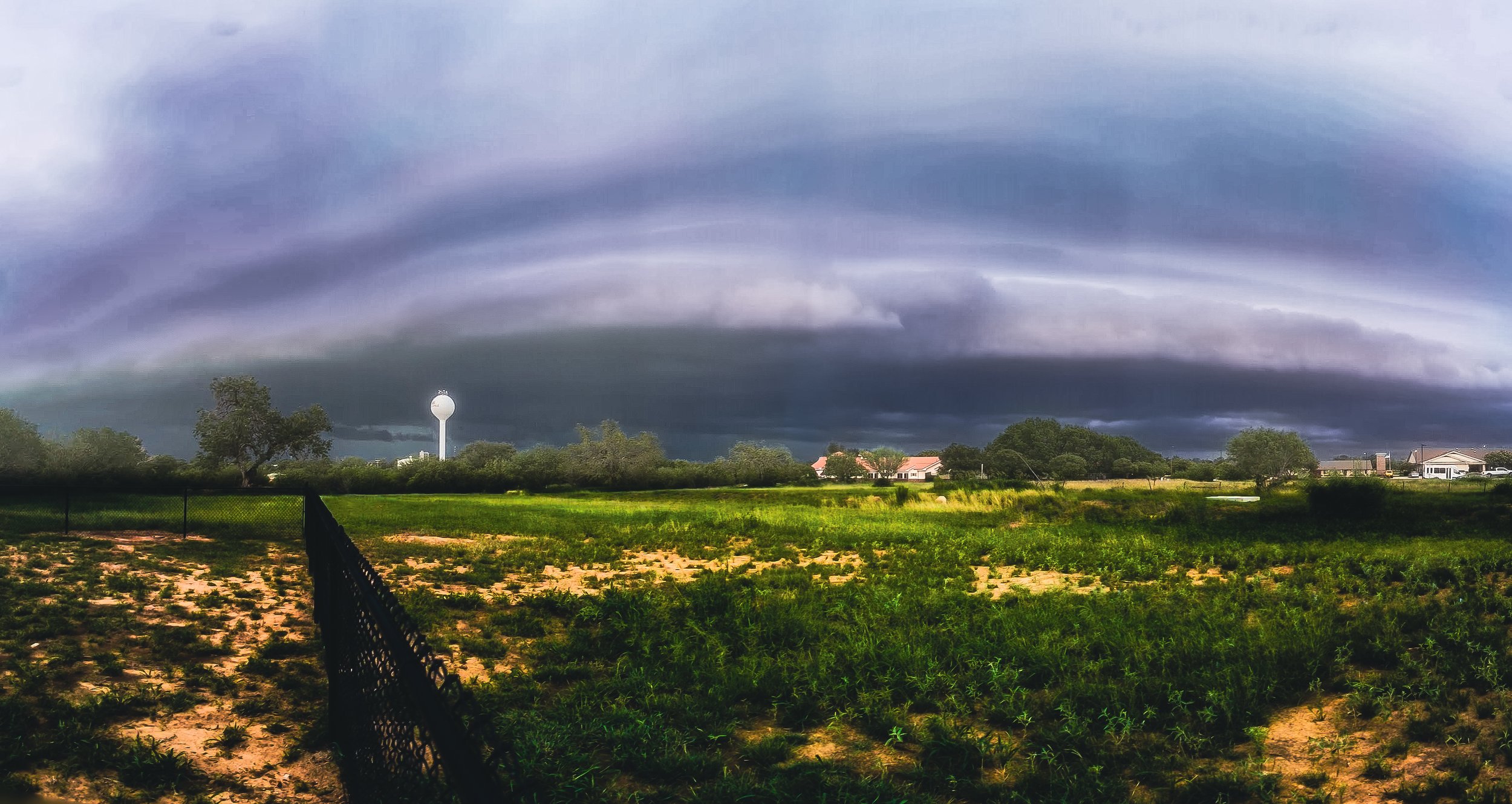 Dark storm clouds looming over a suburban neighborhood with single-story houses, a water tower, and green fields in the foreground.