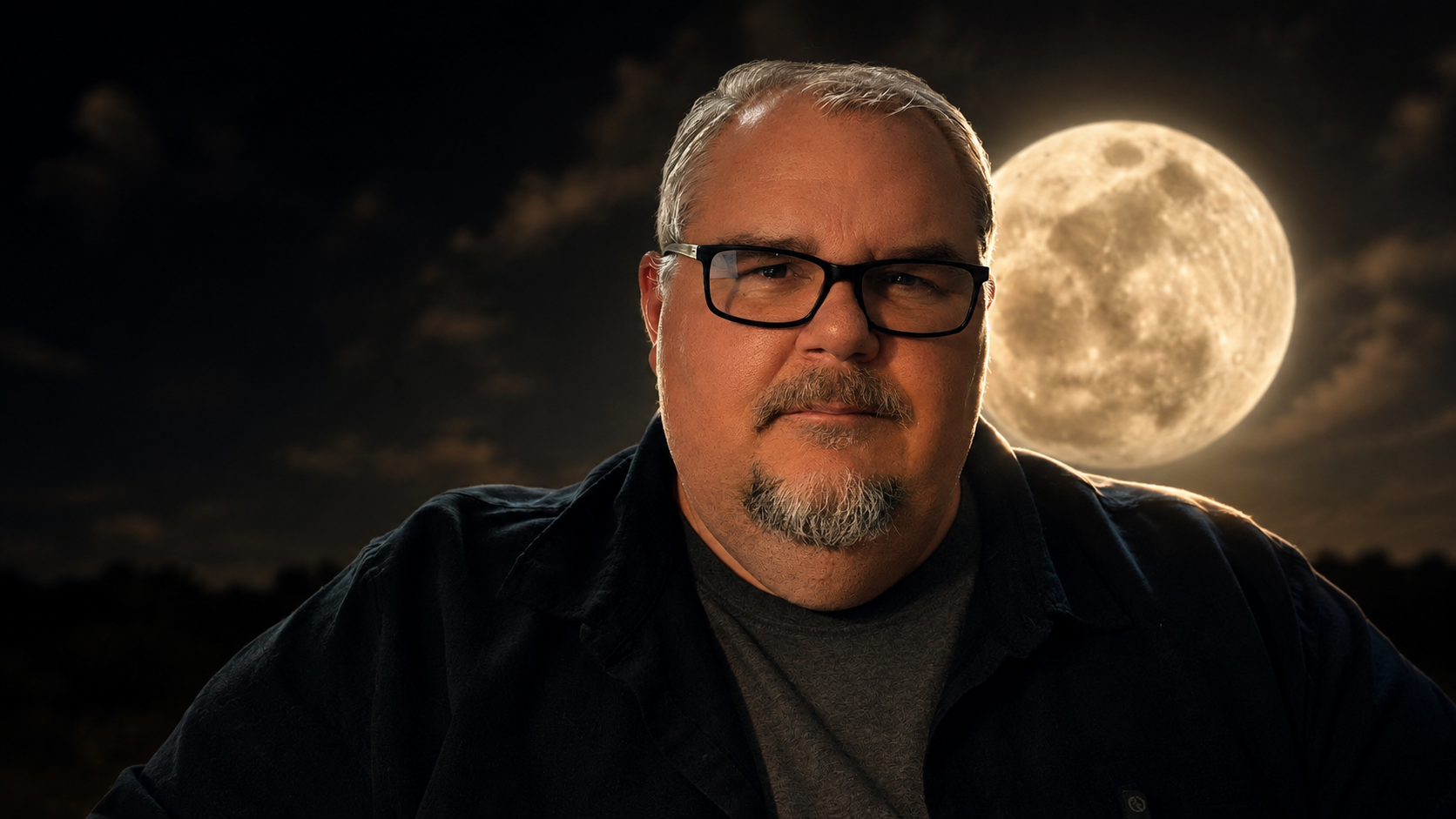A man with glasses and a beard posing outdoors at night with a full moon in the background.