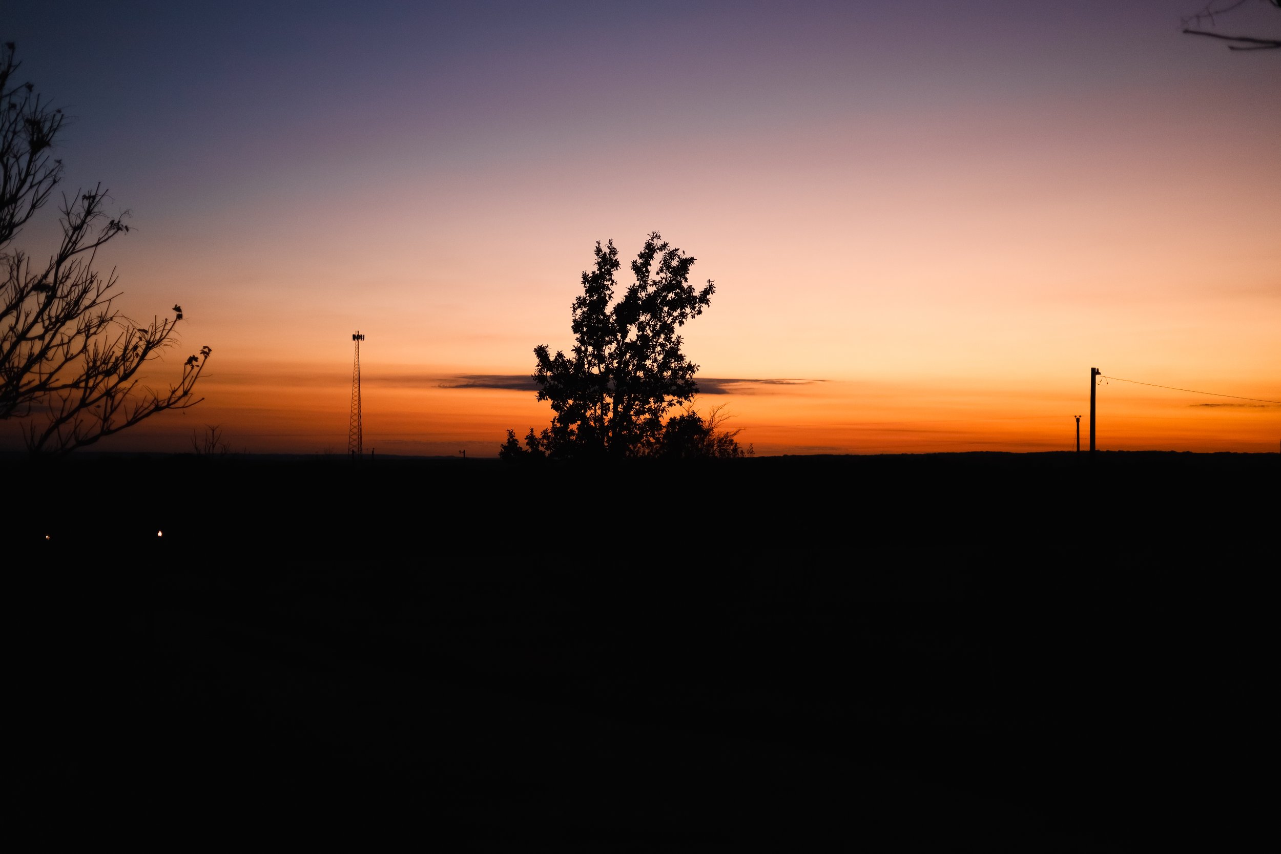 A landscape at dusk with a colorful sunset sky, silhouette of trees, a windmill, and utility poles.
