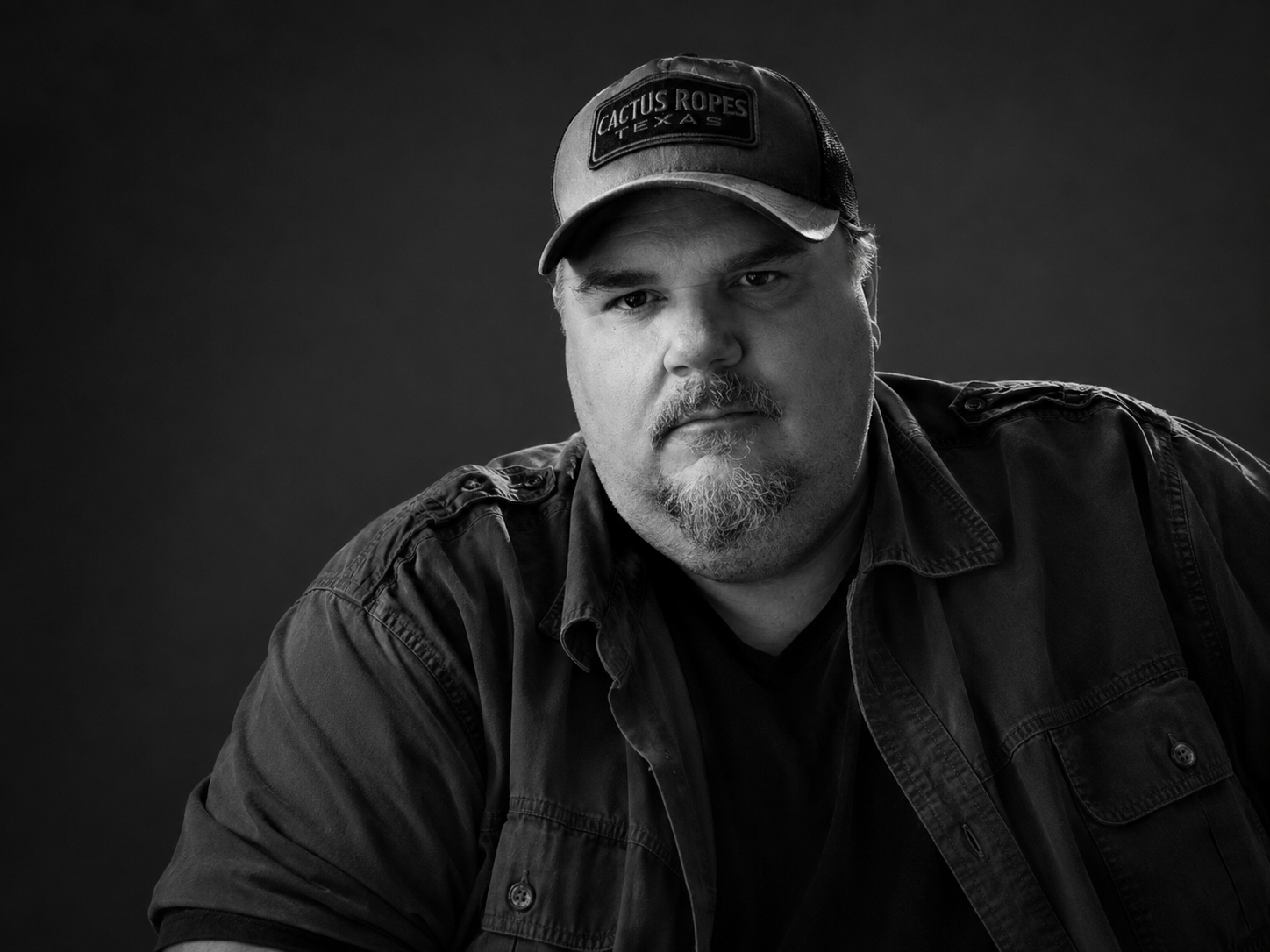 Black and white photo of a man with a beard and mustache wearing a cap that says "Cactus Ropes Texas" and a dark jacket, looking directly at the camera against a dark background.
