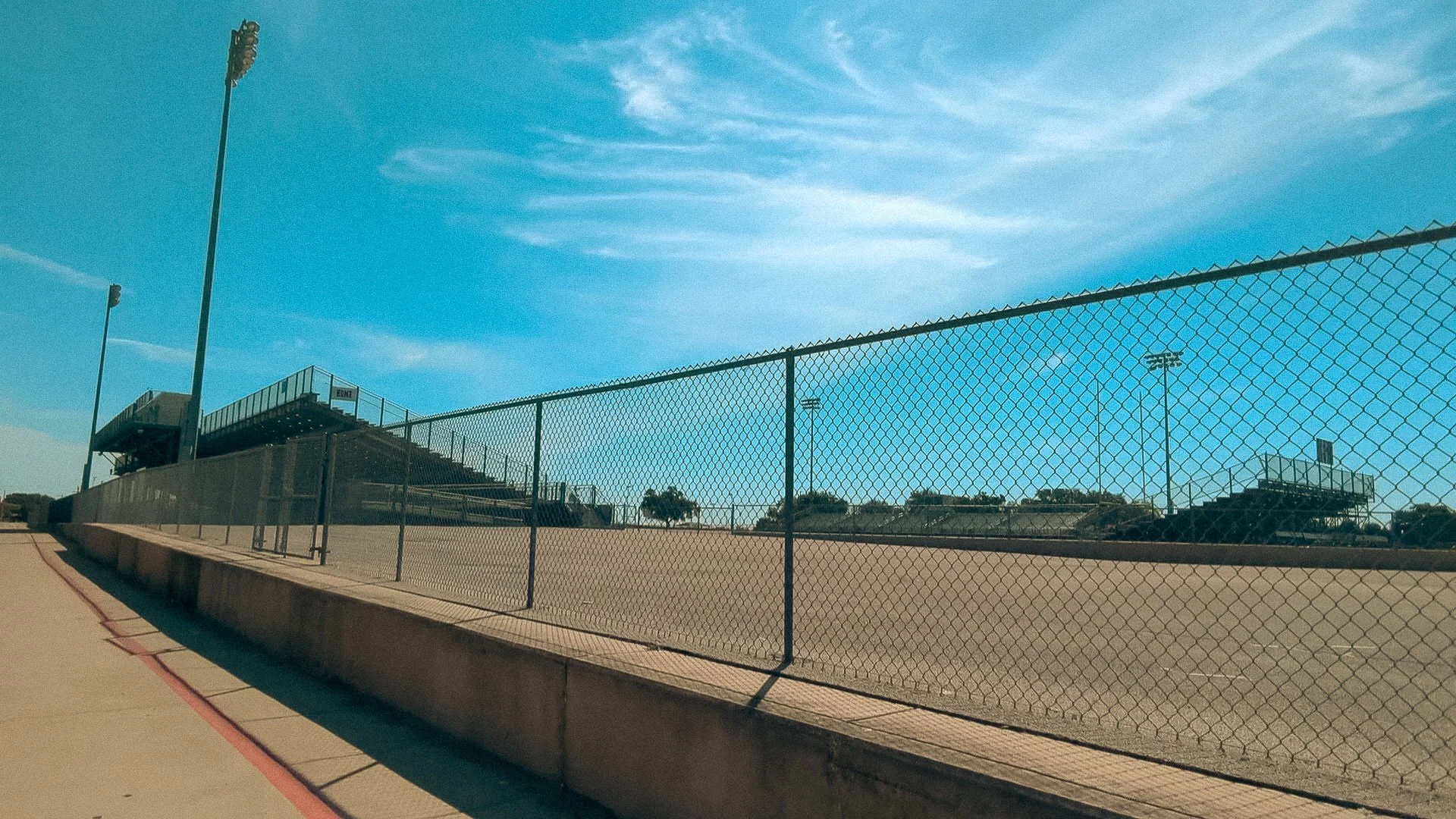 Bleachers and lighting poles at a sports field, enclosed by a chain-link fence, under a partly cloudy sky.