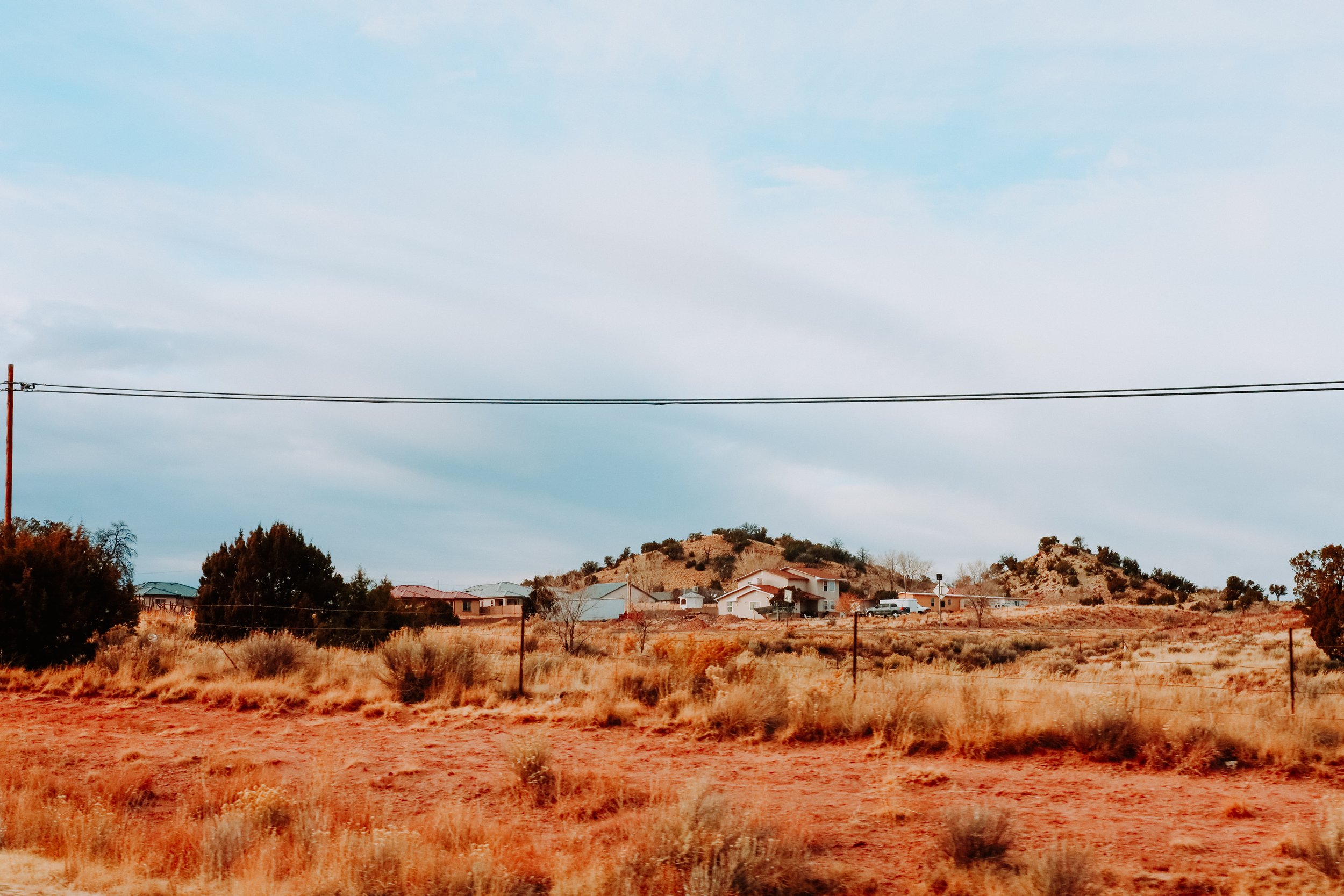 Rural landscape with dry grass, scattered houses, a hill, and power lines under a cloudy sky.