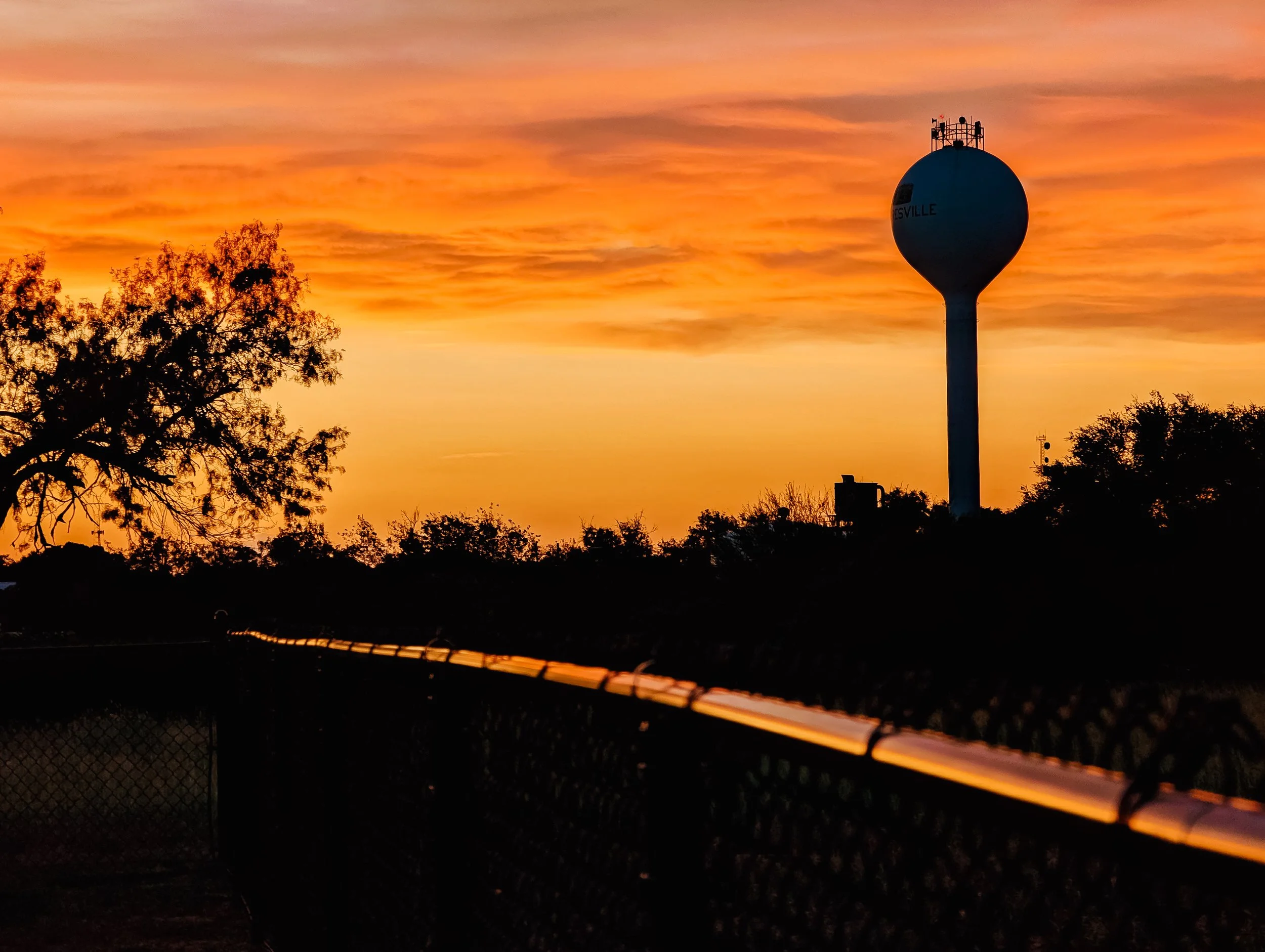 Sunset sky with orange and pink clouds, silhouetted trees, water tower, and a chain-link fence in the foreground.