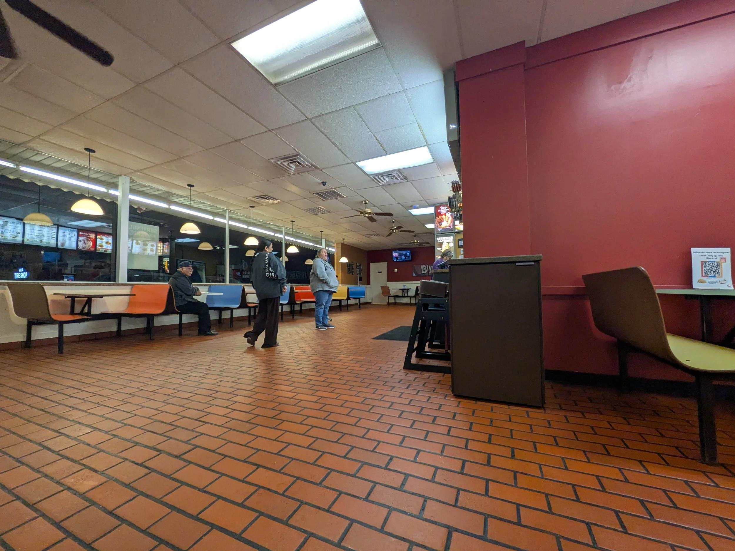 Interior view of a casual restaurant or fast food place with hanging lights, a red accent wall, and a seating area with mostly empty chairs. There are a few people standing and sitting, and digital menu boards are visible behind the counter.