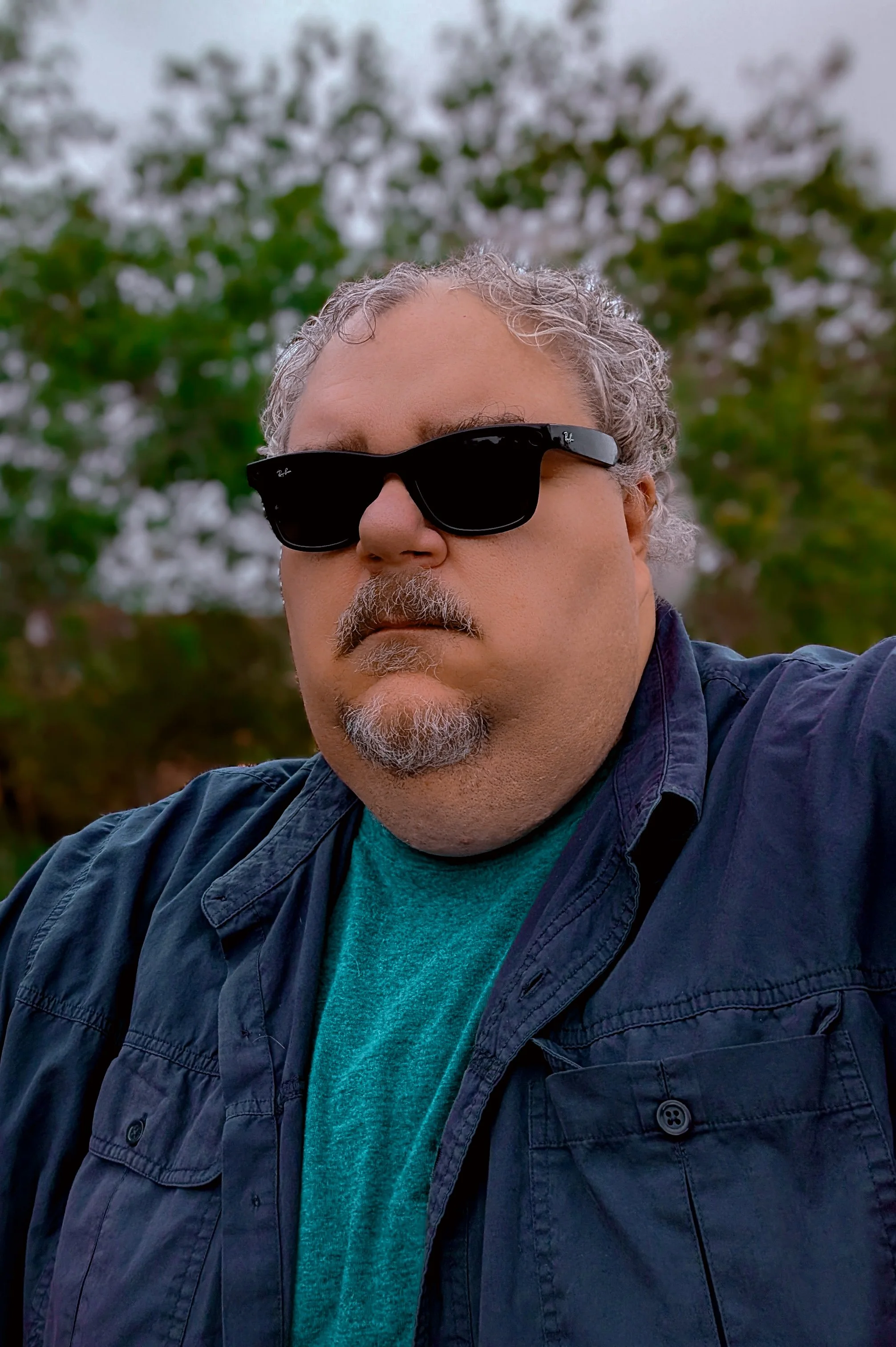 Close-up of a middle-aged man wearing black sunglasses, a green shirt, and a navy blue jacket, outdoors with blurred trees in the background.