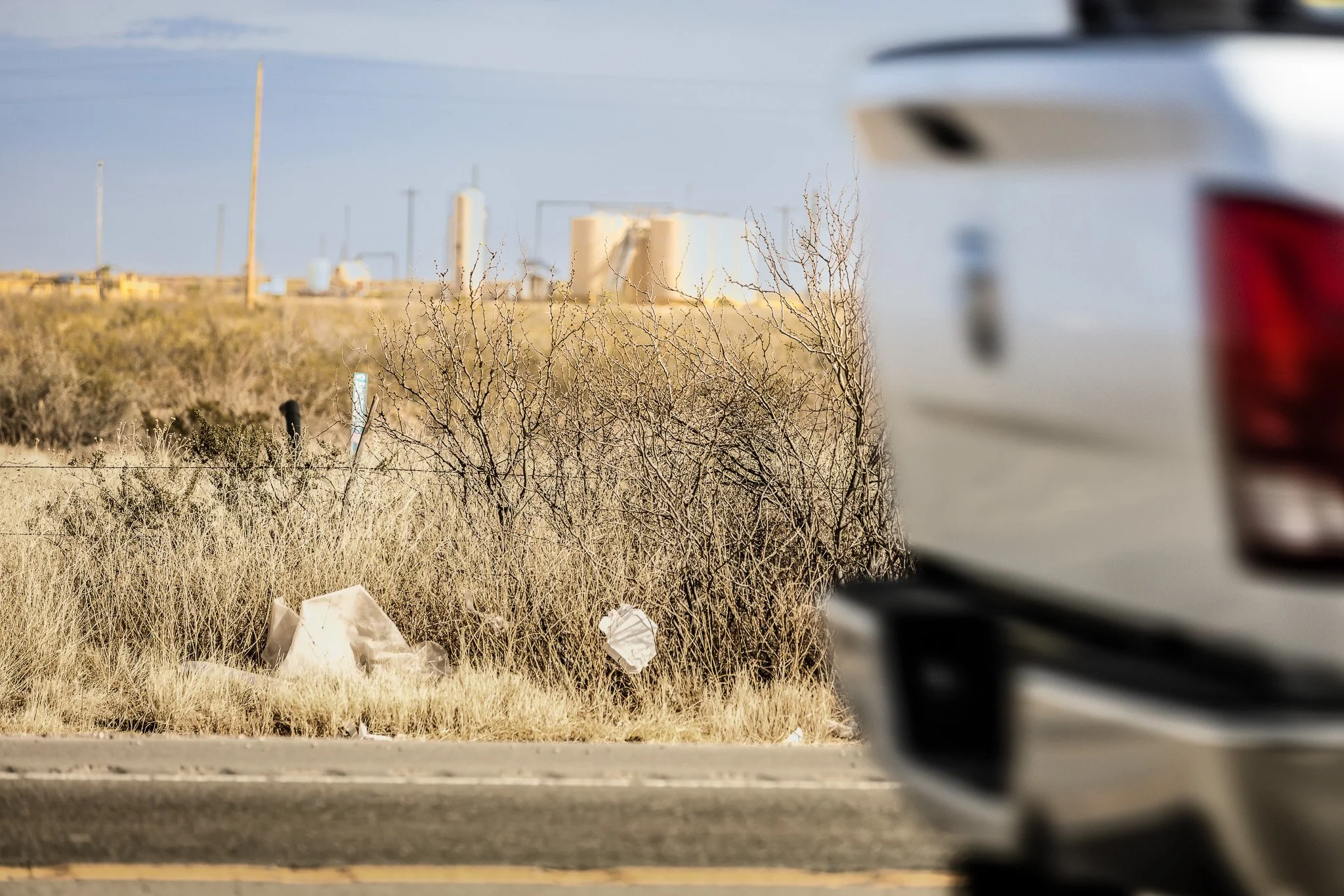 Partially obscured view of a white vehicle on the right side of the image, with a barren, grassy area and dry shrubs in the background. There are some white trash bags or paper on the ground. The background includes industrial structures with tanks and buildings, under a clear sky.
