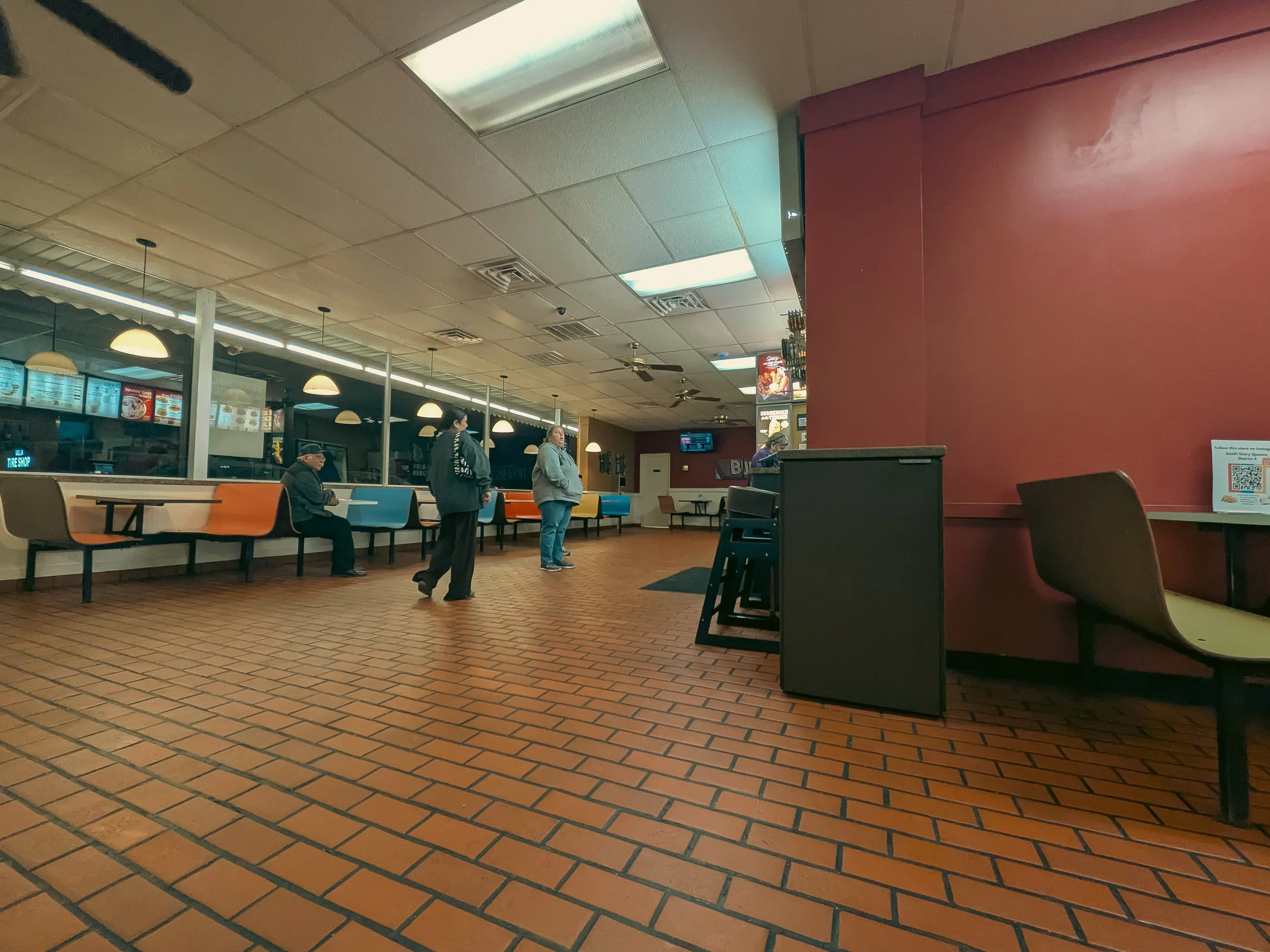 Inside a fast food restaurant with orange-tiled flooring, a red wall on the right, and a line of chairs. Several people are standing or sitting, waiting or eating. The menu boards are visible behind the counter, and hanging lamps illuminate the space.