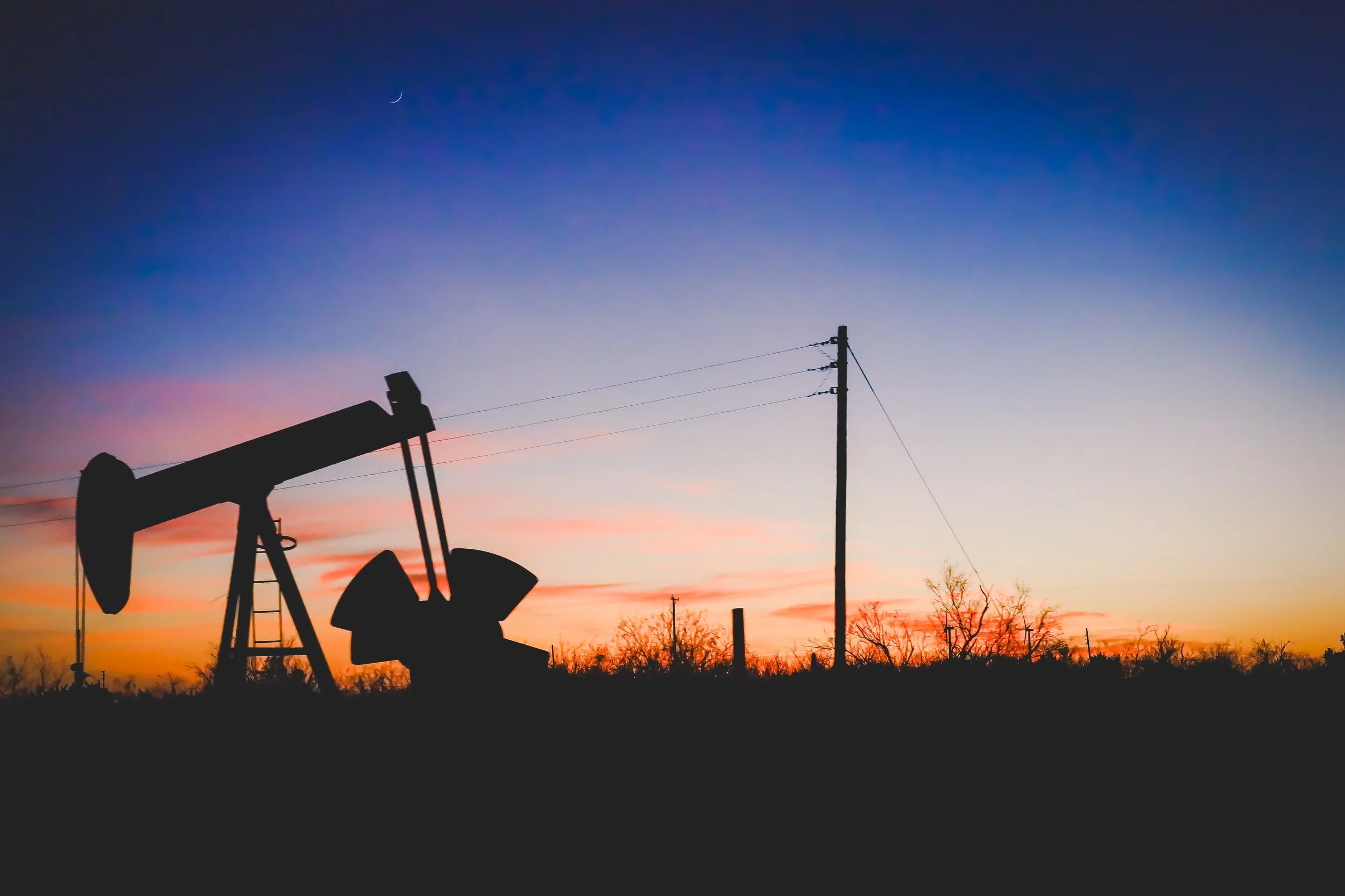 Silhouettes of oil pumpjack and utility pole against a colorful sunset sky with a small crescent moon.