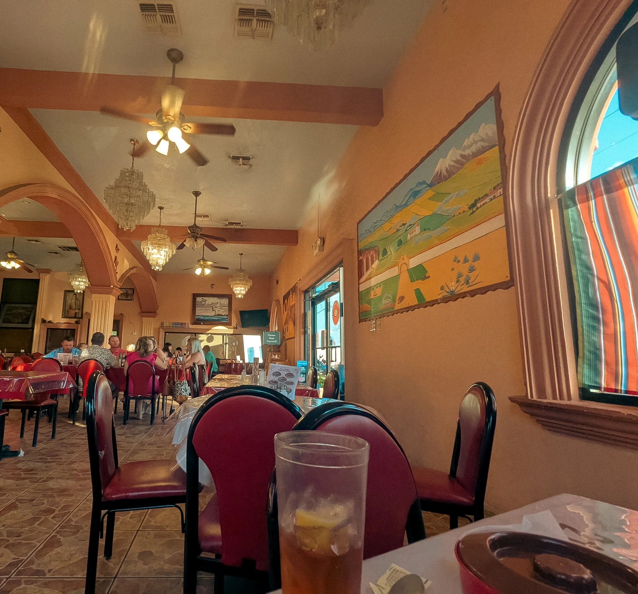 Interior of a restaurant or diner with tables, red chairs, and multiple ceiling lights and fans. A colorful landscape painting is on the wall near a window with striped curtains. Several people are seated at tables.