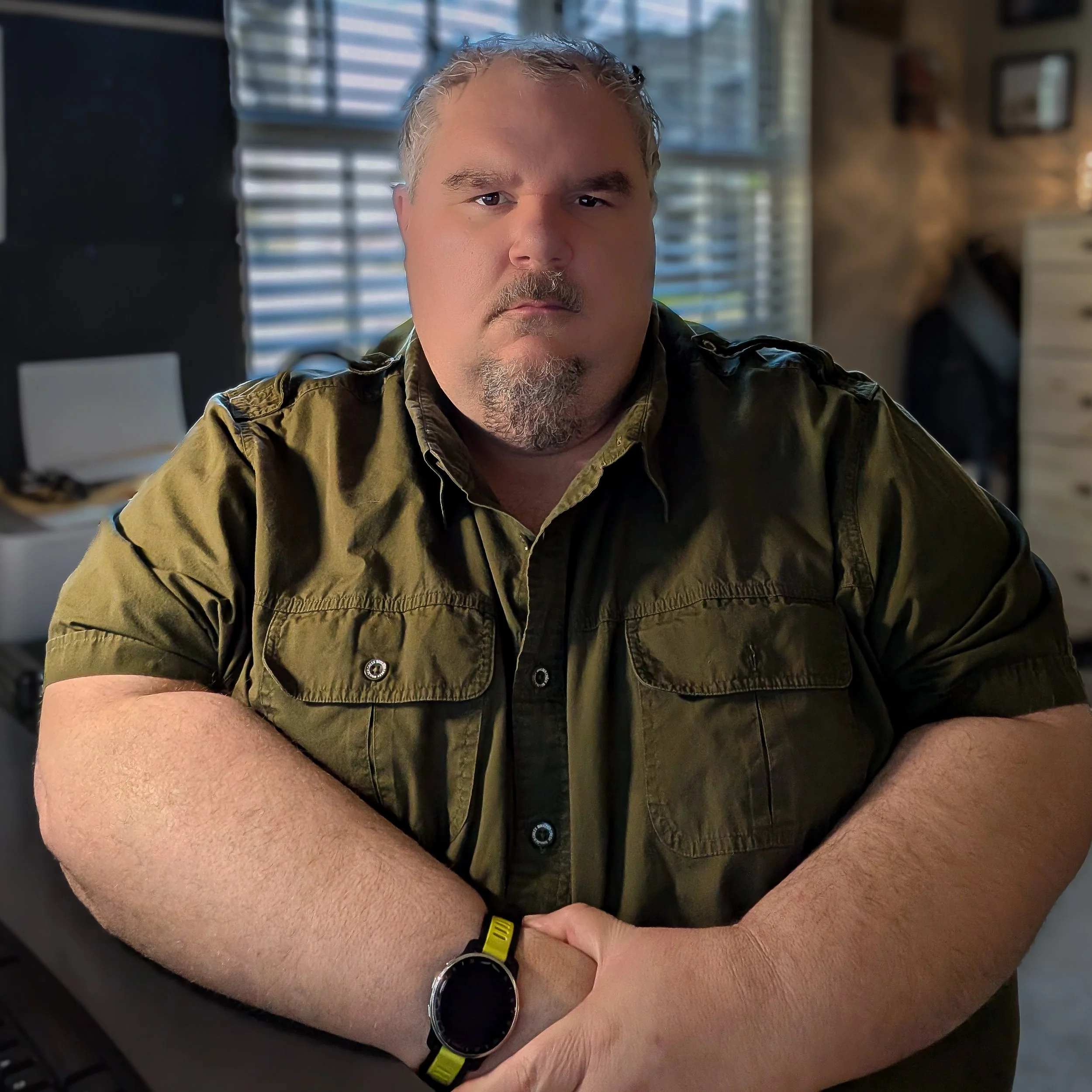 A man with gray hair and a beard wearing a green shirt, a watch, and a yellow wristband sitting with his arms crossed. The background includes a window with blinds and some picture frames on the wall.