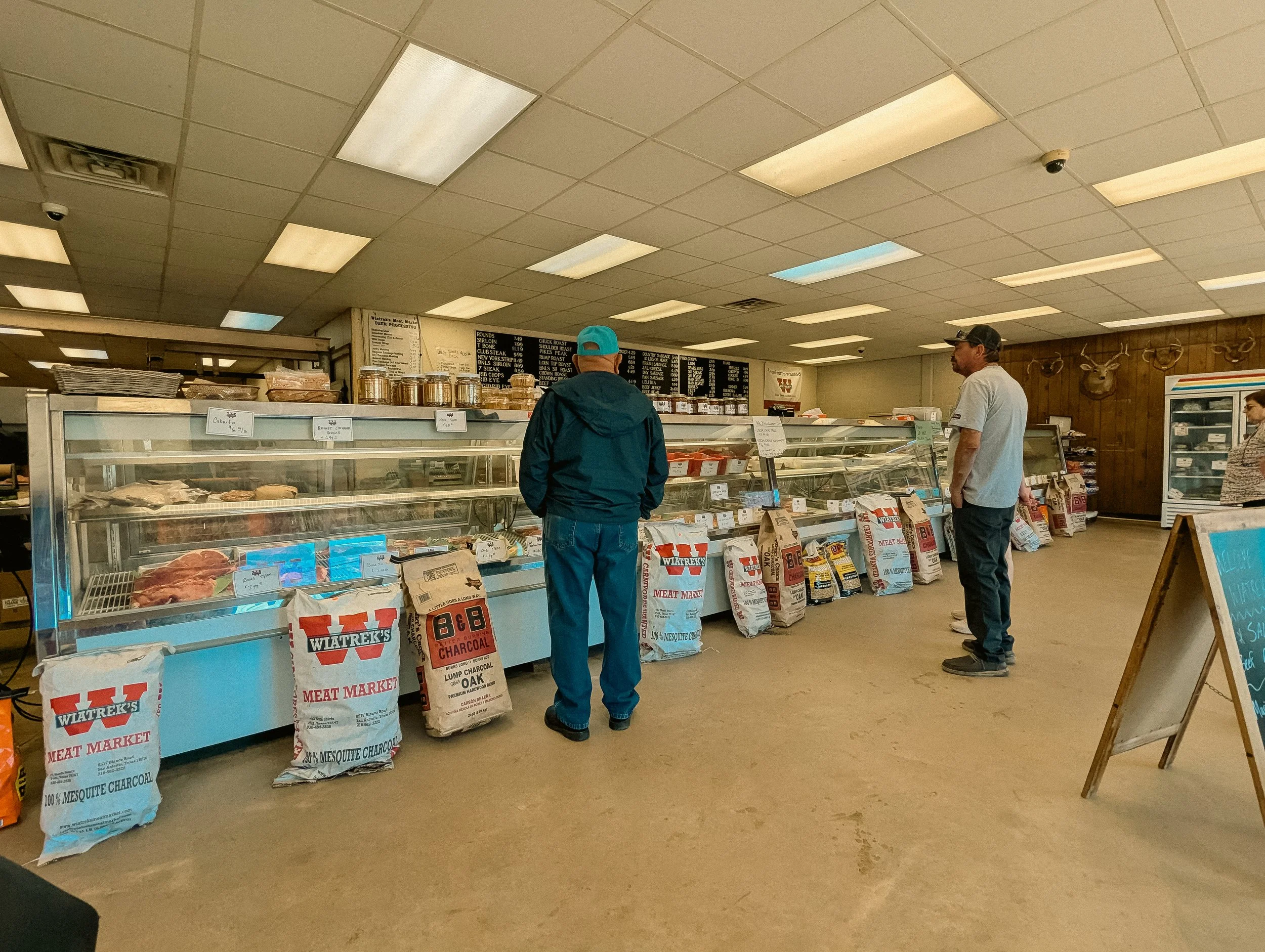 Inside a meat market with two men looking at meat and a woman behind the counter, with bags of charcoal and grocery items visible.