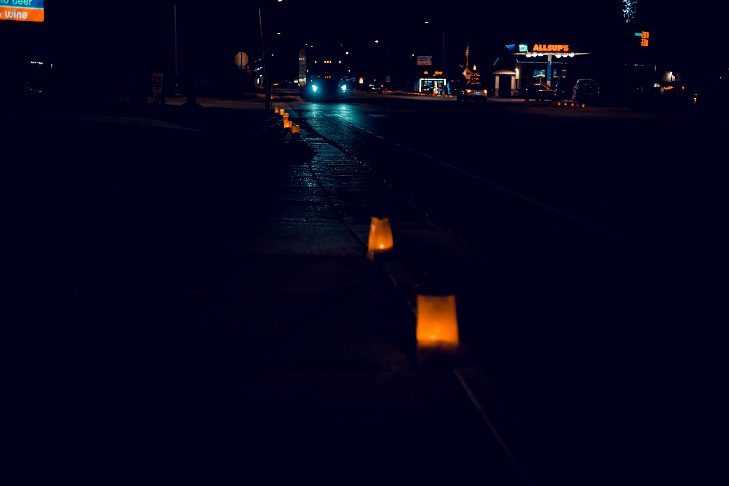 Night street scene with luminaries lining the sidewalk, car headlights in the distance, and a gas station in the background.