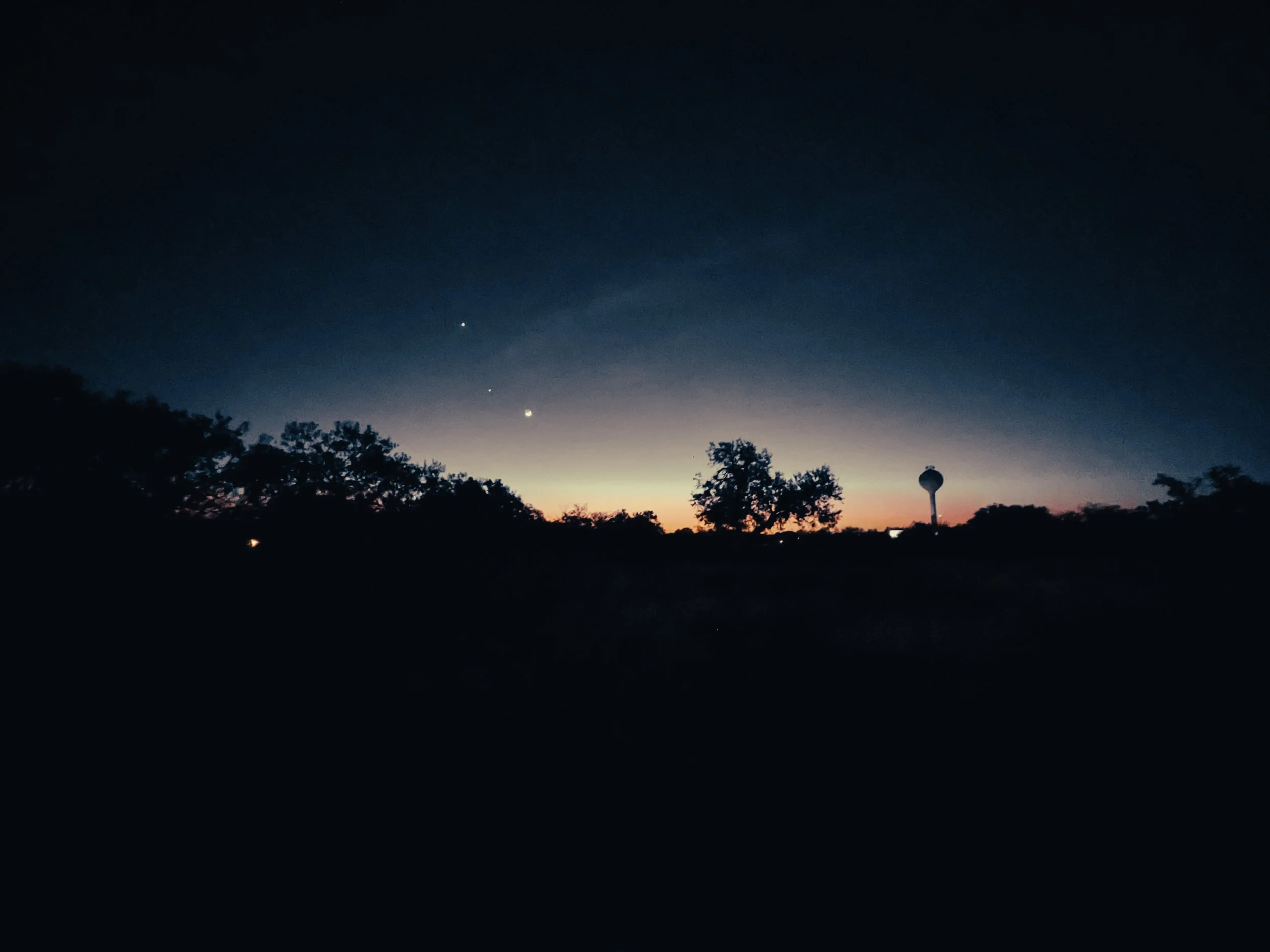 Nighttime sky with the transition from sunset at horizon, silhouettes of trees, and celestial bodies including planets and stars visible in the sky.