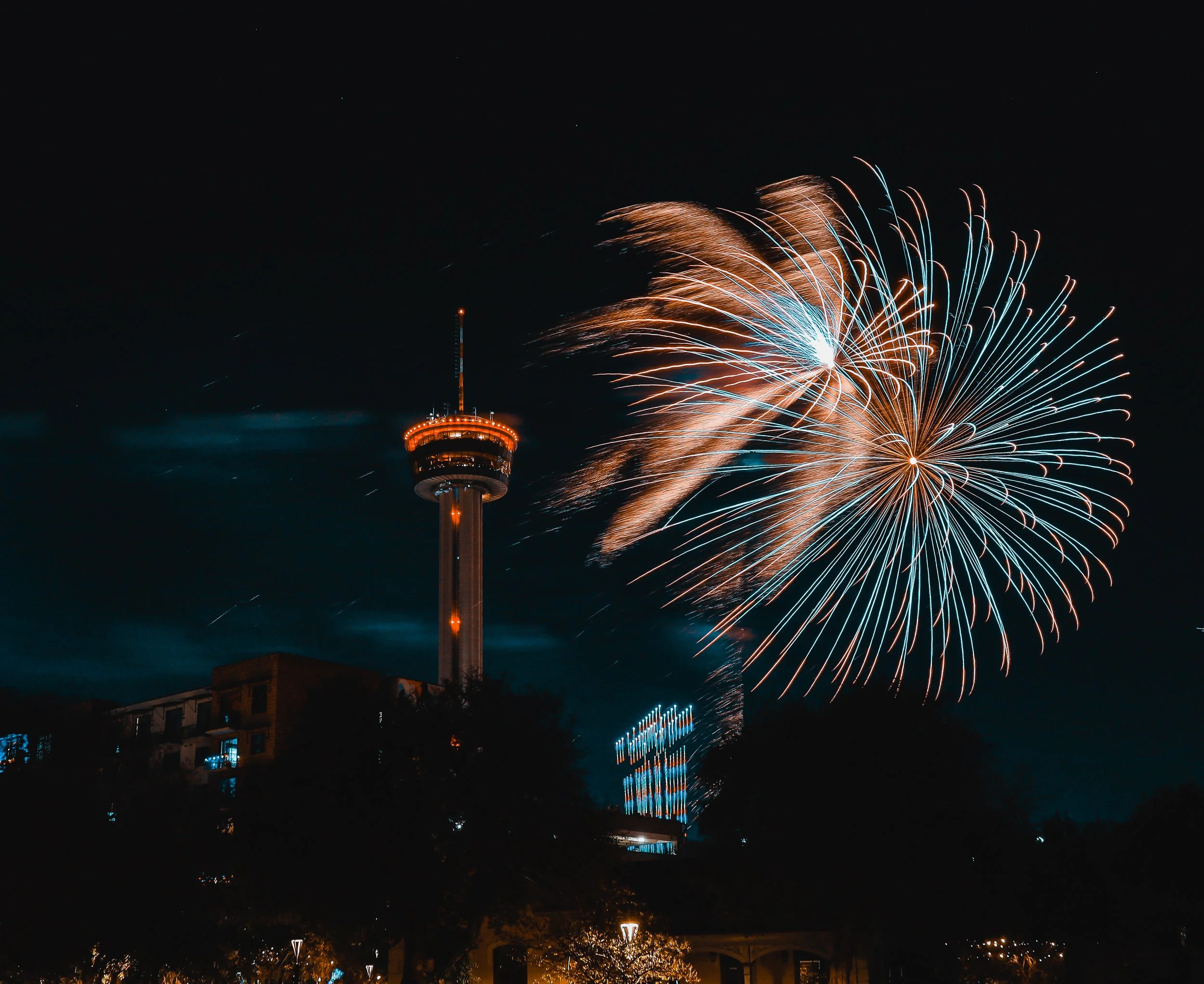 Fireworks bursting in the night sky near the Tower of the Americas in San Antonio, with city lights and silhouettes of trees below, capturing a hopeful New Year’s celebration.