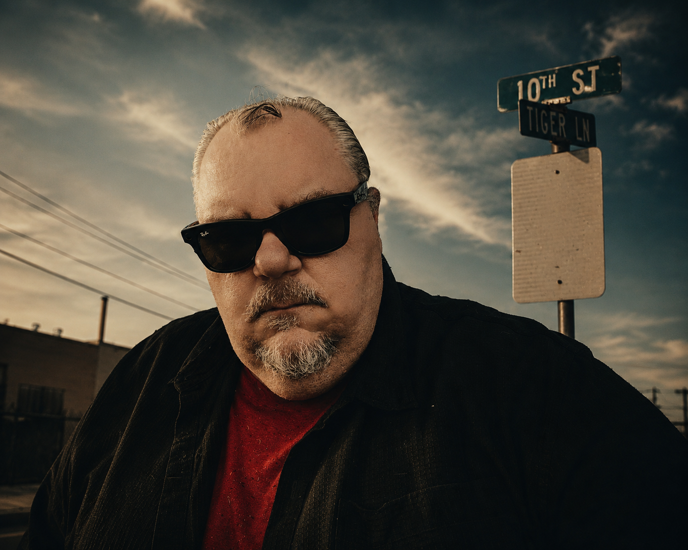 A man with a goatee and glasses, wearing a black jacket and red shirt, standing outdoors during sunset with street signs for 10th Street and Tiger Lane in the background.