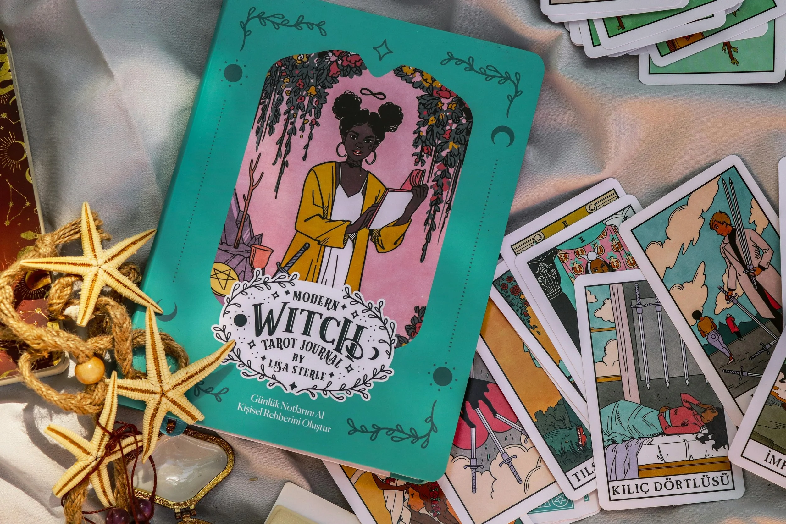 A tarot journal titled 'Modern Witch' by Lisa Sterle, featuring a woman with dark hair styled in two buns, reading a book against a pink background with hanging vines. Surrounding it are tarot cards laid out on a silver surface, with some cards displayed revealing illustrations of swords and characters. There are also starfish and shell decorations on the left side of the image.