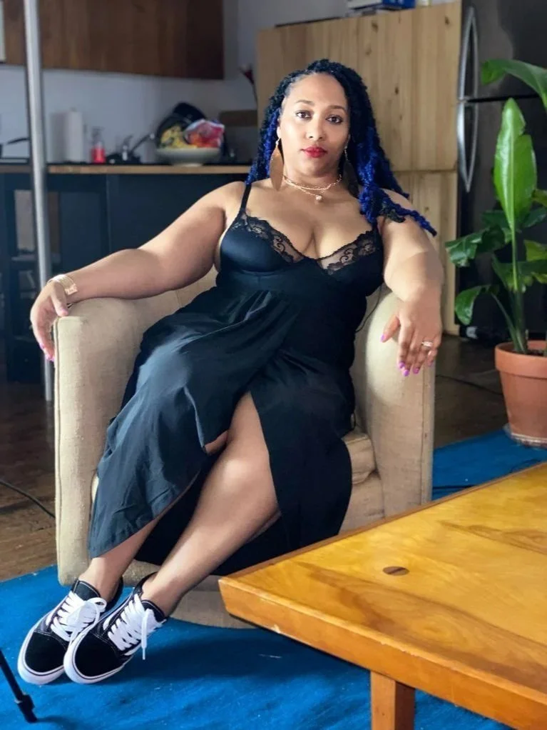 A woman with blue braided hair sitting on a beige armchair in a room with wooden furniture, wearing a black lace dress and sneakers.