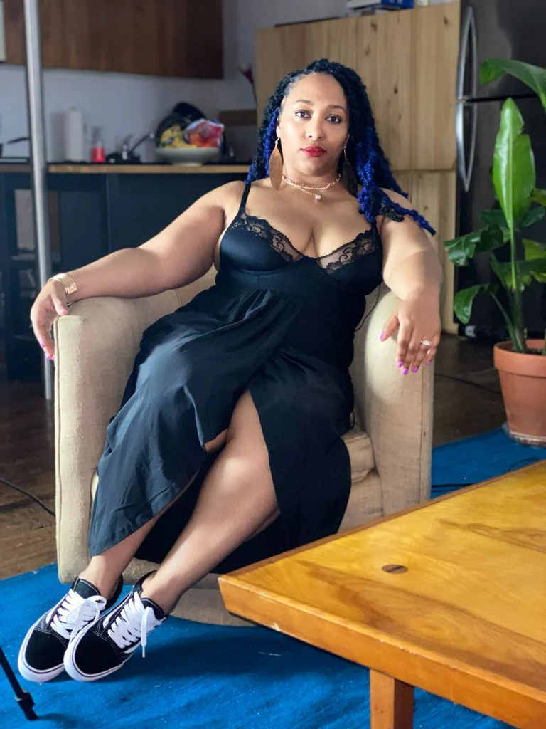 Woman sitting on a beige armchair in a kitchen, wearing a black dress with lace details and sneakers, with blue braids hairstyle, gold jewelry, and a confident expression.
