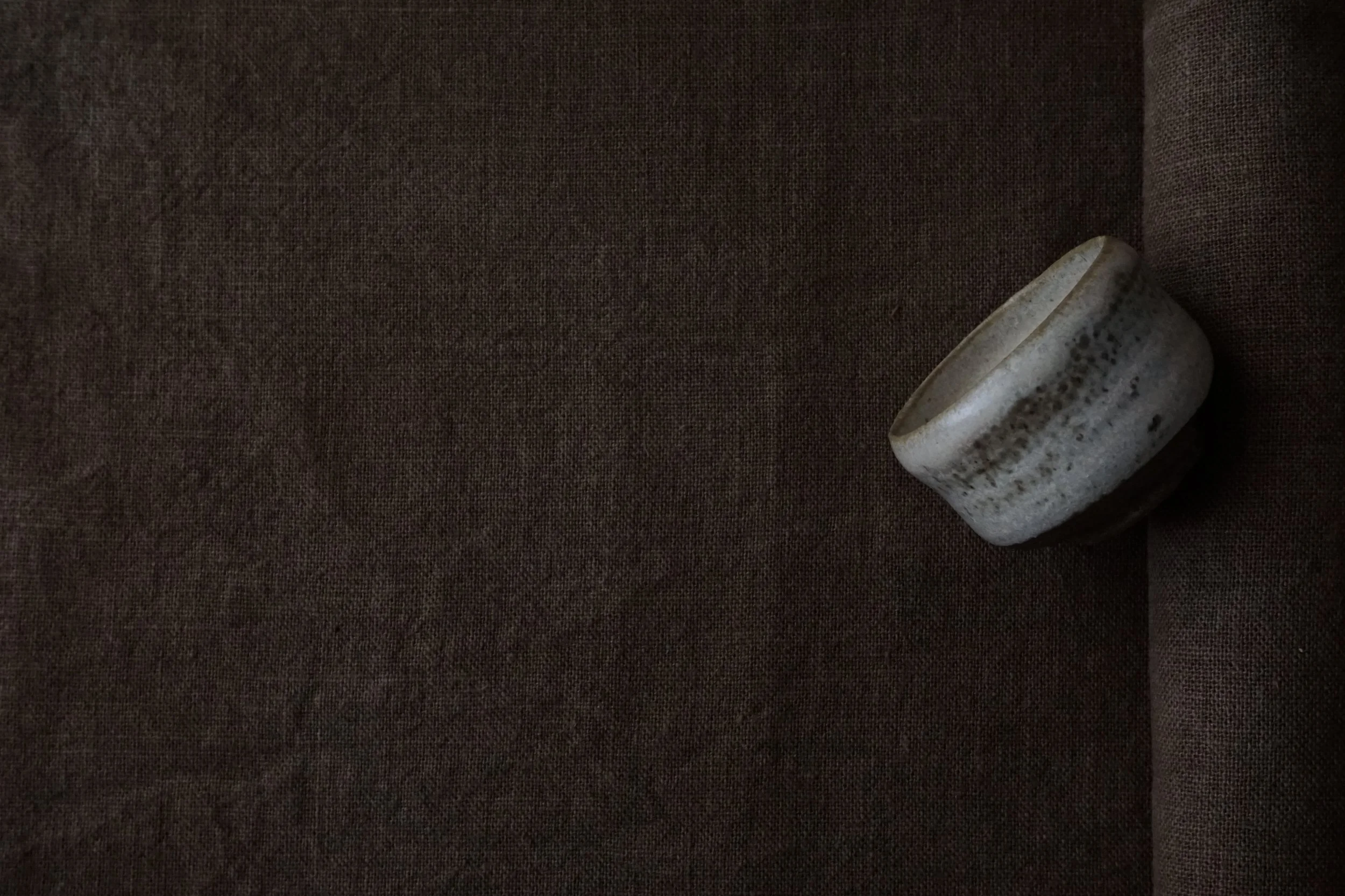 Petite tasse ronde en céramique beige avec tachettes brunes, posée sur un fond en tissu brun foncé.