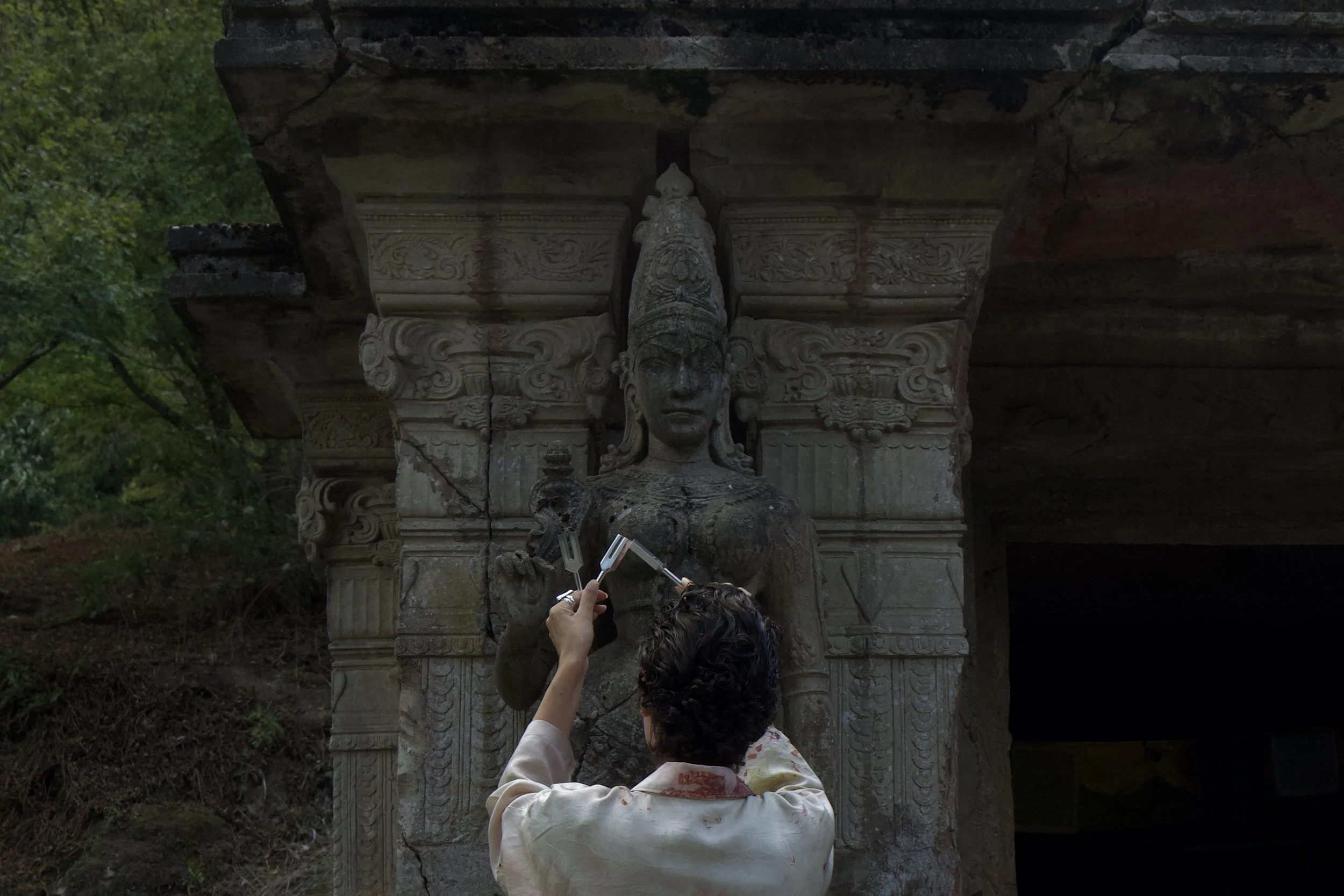 Une personne prenant une photo d'une statue ancienne en pierre représentant une déesse hindoue, située sous une structure rocheuse dans un environnement naturel.