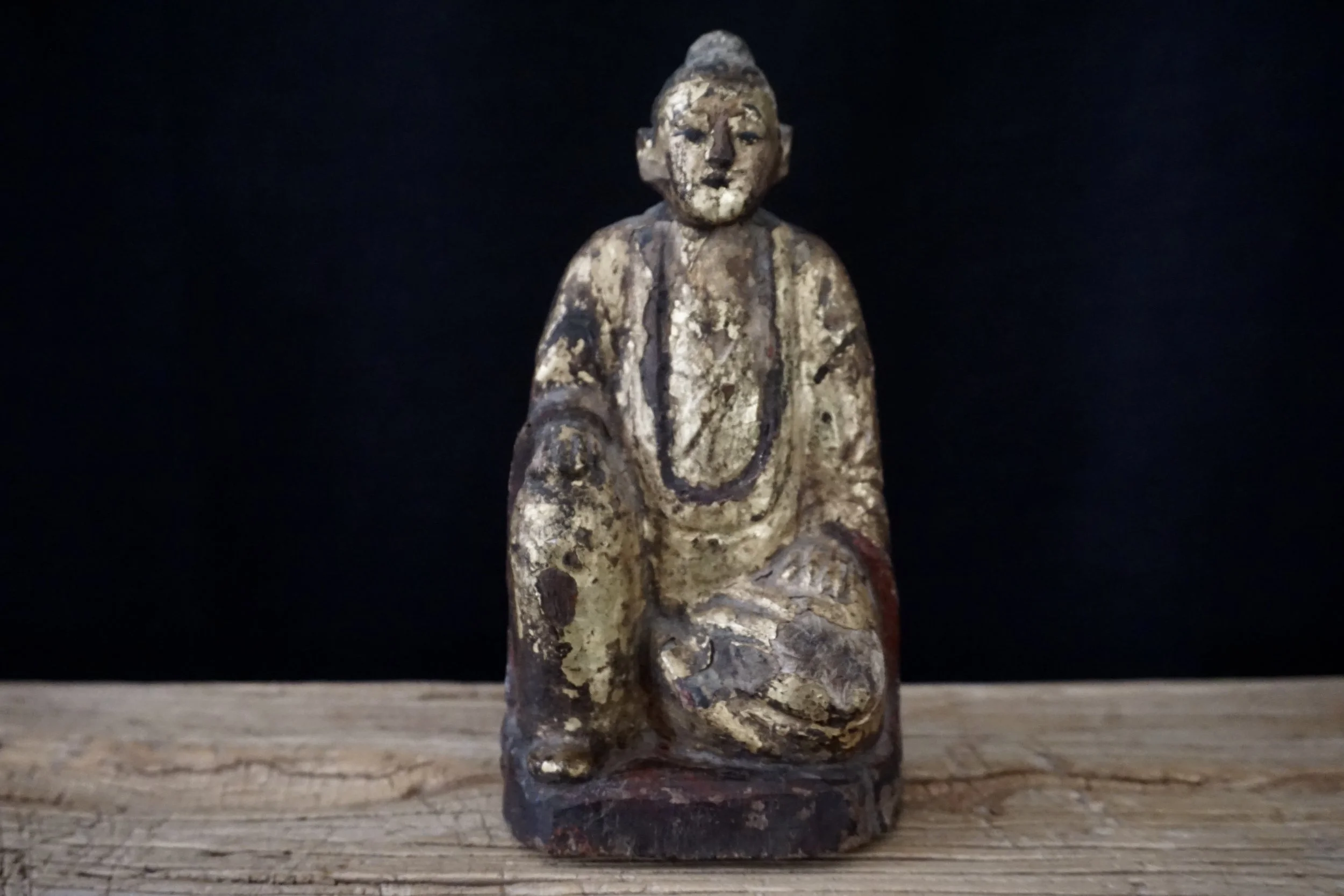 Statue votive antique en bois représentant un ancêtre Taoïste assis avec un objet dans les mains, sur une surface en bois, fond noir. Wabi sabi, laque et or.