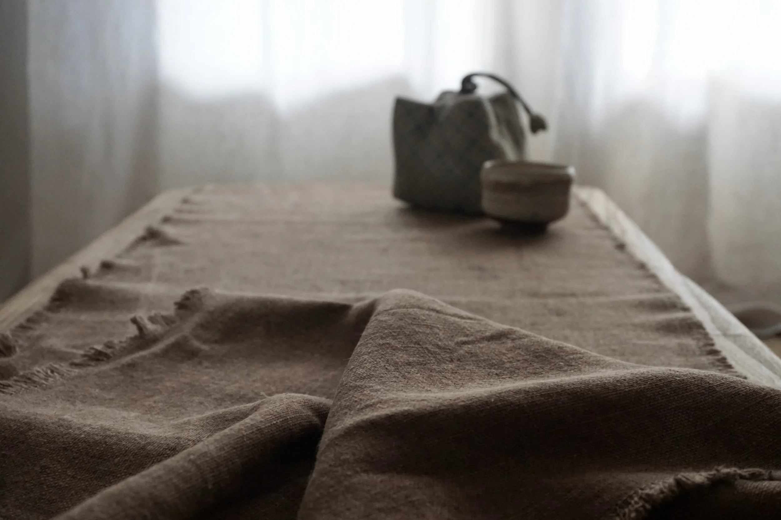 Une nappe en lin beige posée sur une table en bois, avec un sac en tissu et un petit objet en bois ou céramique en arrière-plan, dans une pièce avec lumière naturelle