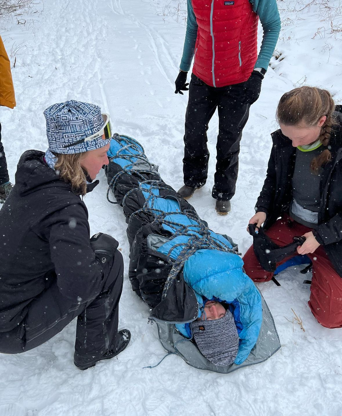 A group of people in winter clothing assisting a person lying on the snow inside a rescue net.