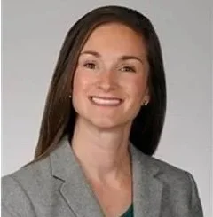 Professional woman with long brown hair wearing a gray blazer, smiling against a plain background.