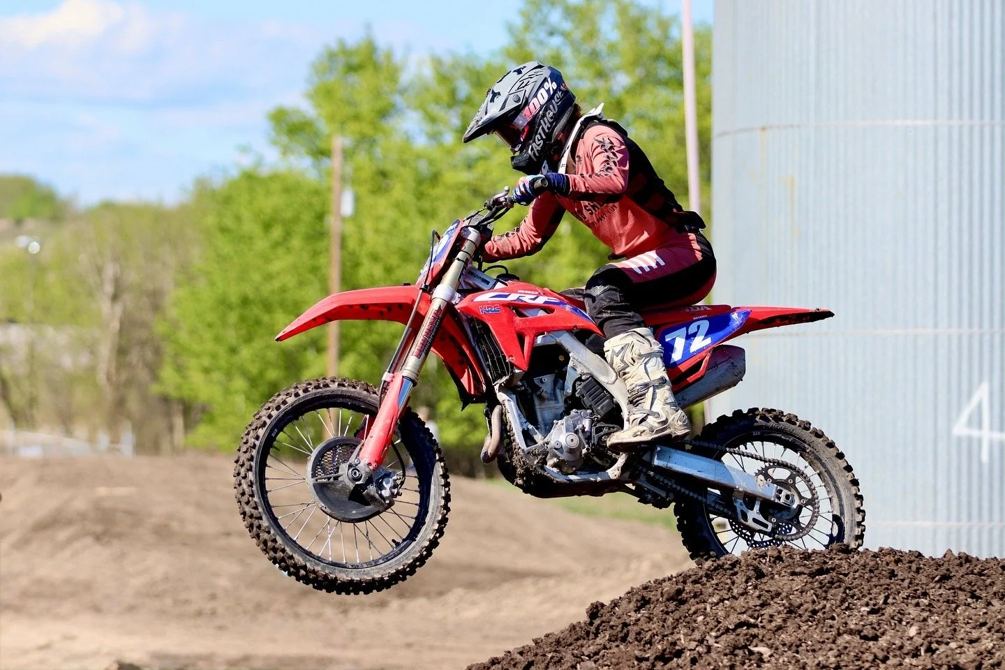 A person riding a red dirt bike over a mound of dirt during daytime, wearing a helmet and protective gear.