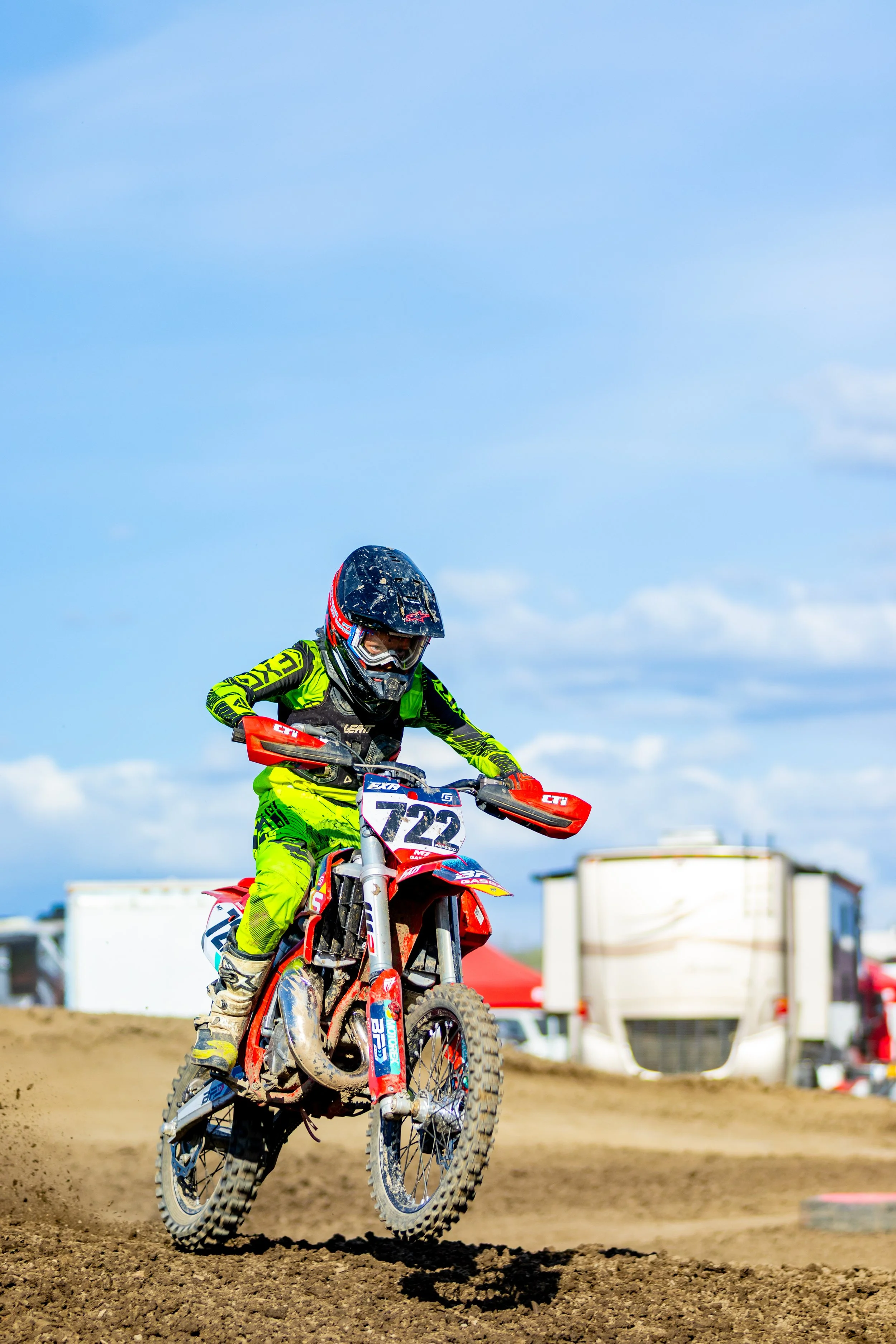 Motocross rider in bright green gear riding a dirt bike on a dirt track with a blue sky and scattered clouds in the background.