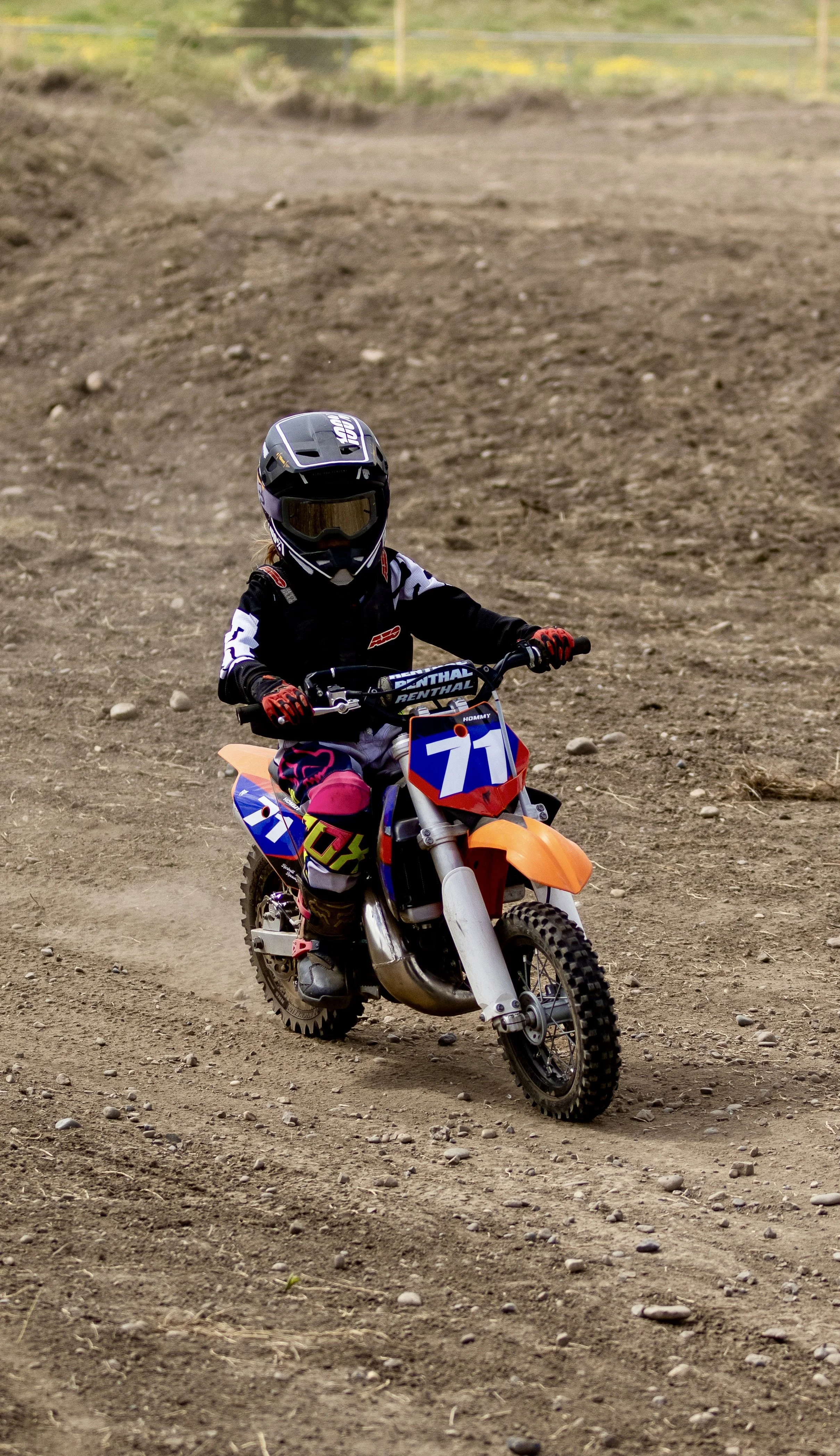 A young motocross rider in black helmet and pink gear riding an orange dirt bike on a dirt track.