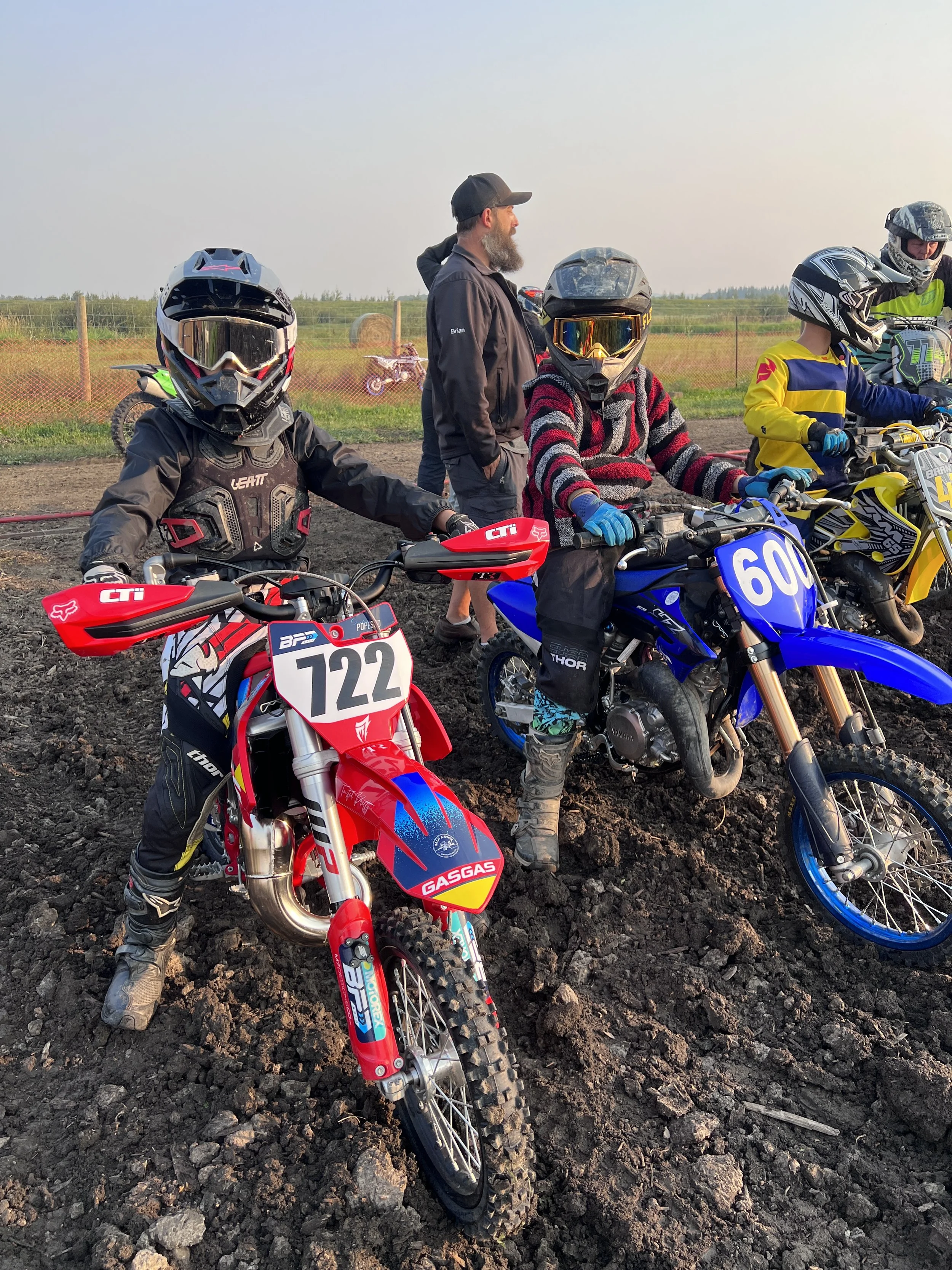 Group of children and an adult on dirt bikes on a dirt track, wearing helmets and protective gear, with a rural landscape in the background.