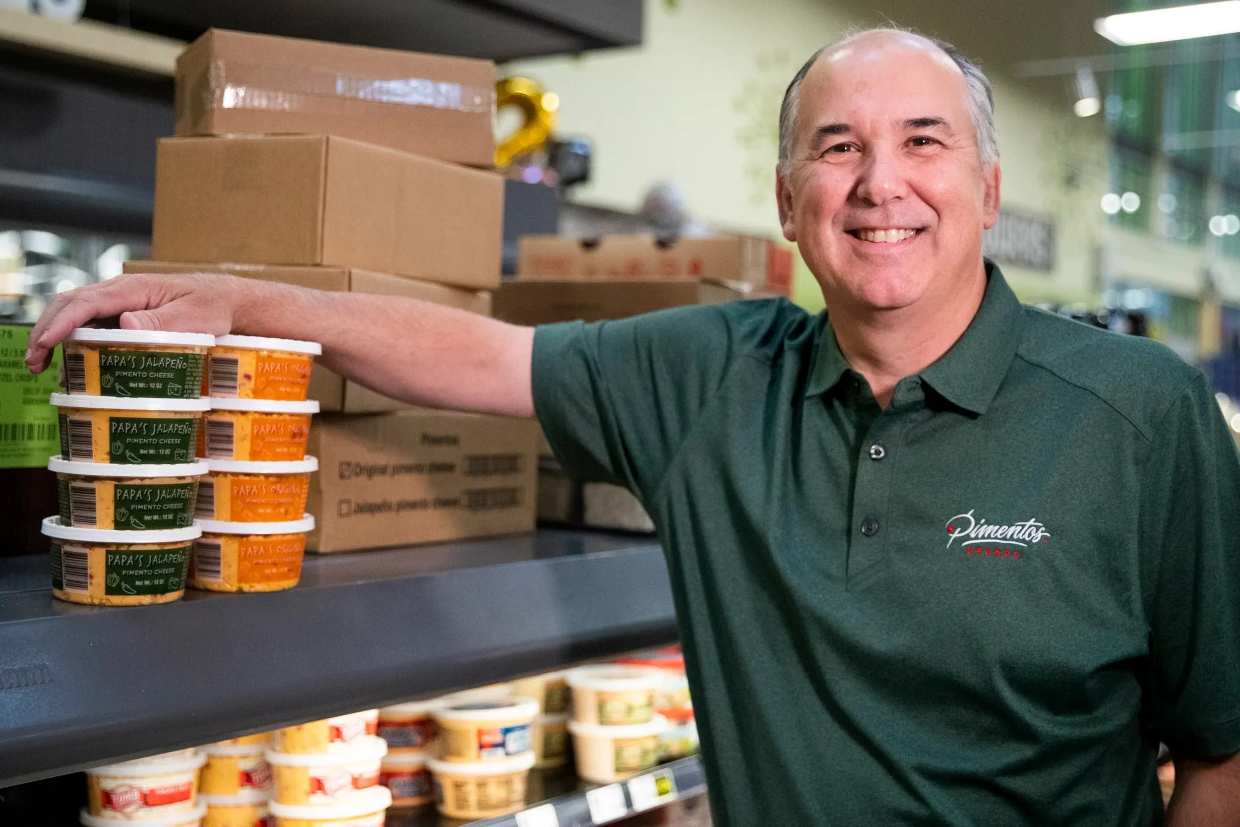 A smiling man in a green shirt standing in a grocery store aisle, with shelves of packaged food products, Specifically, he has his hand resting on a stack of small containers labeled 'Papa's Jalapeño Pimento Cheese'.