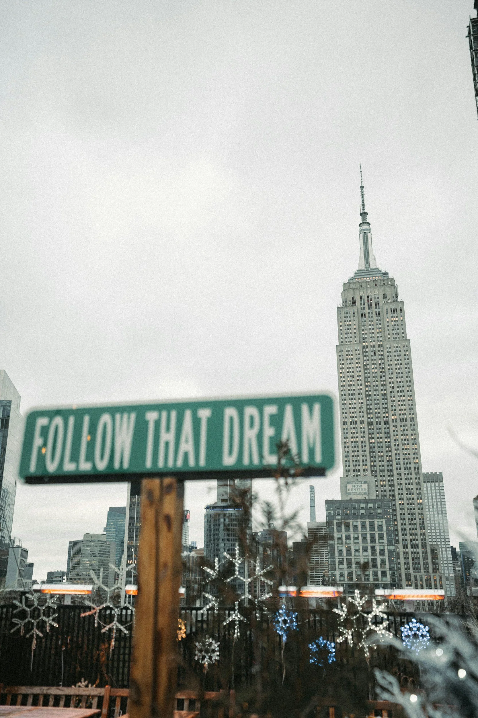 New York City skyline with the Empire State Building in the background and a blurred green sign that says 'Follow That Dream' in the foreground, along with holiday decorations and snowflake lights.