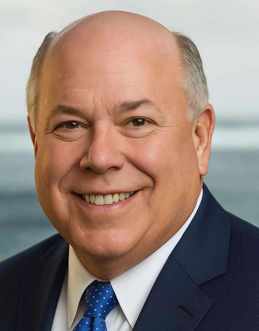 Close-up of a smiling middle-aged man with a bald head, wearing a dark suit, white shirt, and blue polka dot tie, with a blurred background of water and sky.