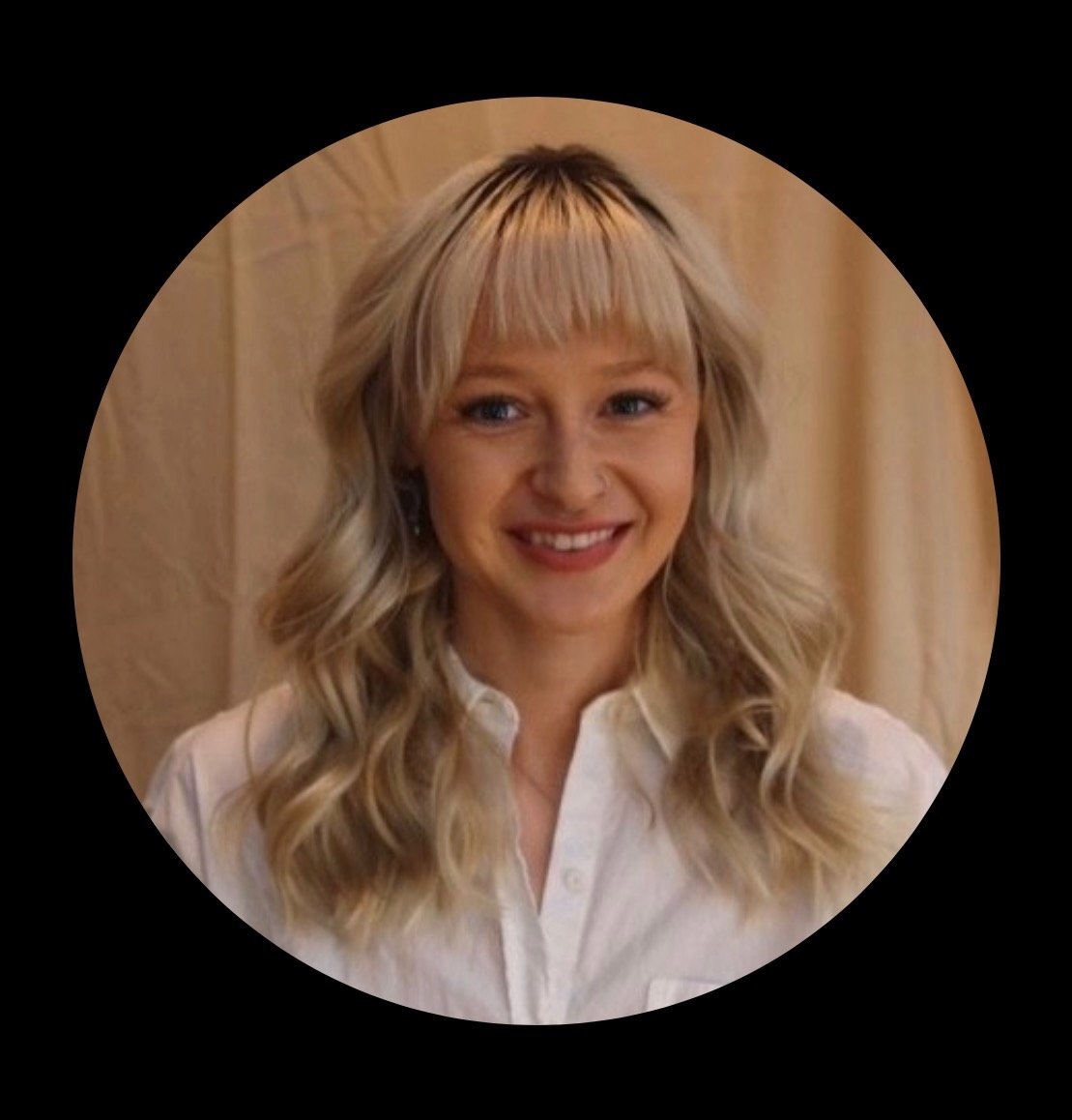 A woman with blonde wavy hair and a white shirt smiling indoors.