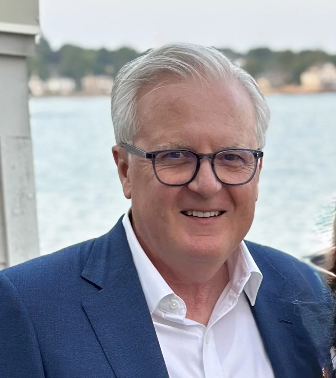 A smiling middle-aged man with gray hair and glasses, wearing a navy suit and white shirt, standing outdoors near a body of water with houses in the background.