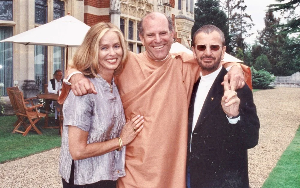 Craig in an orange robe with his arms around two smiling friends in front of a stately brick manor