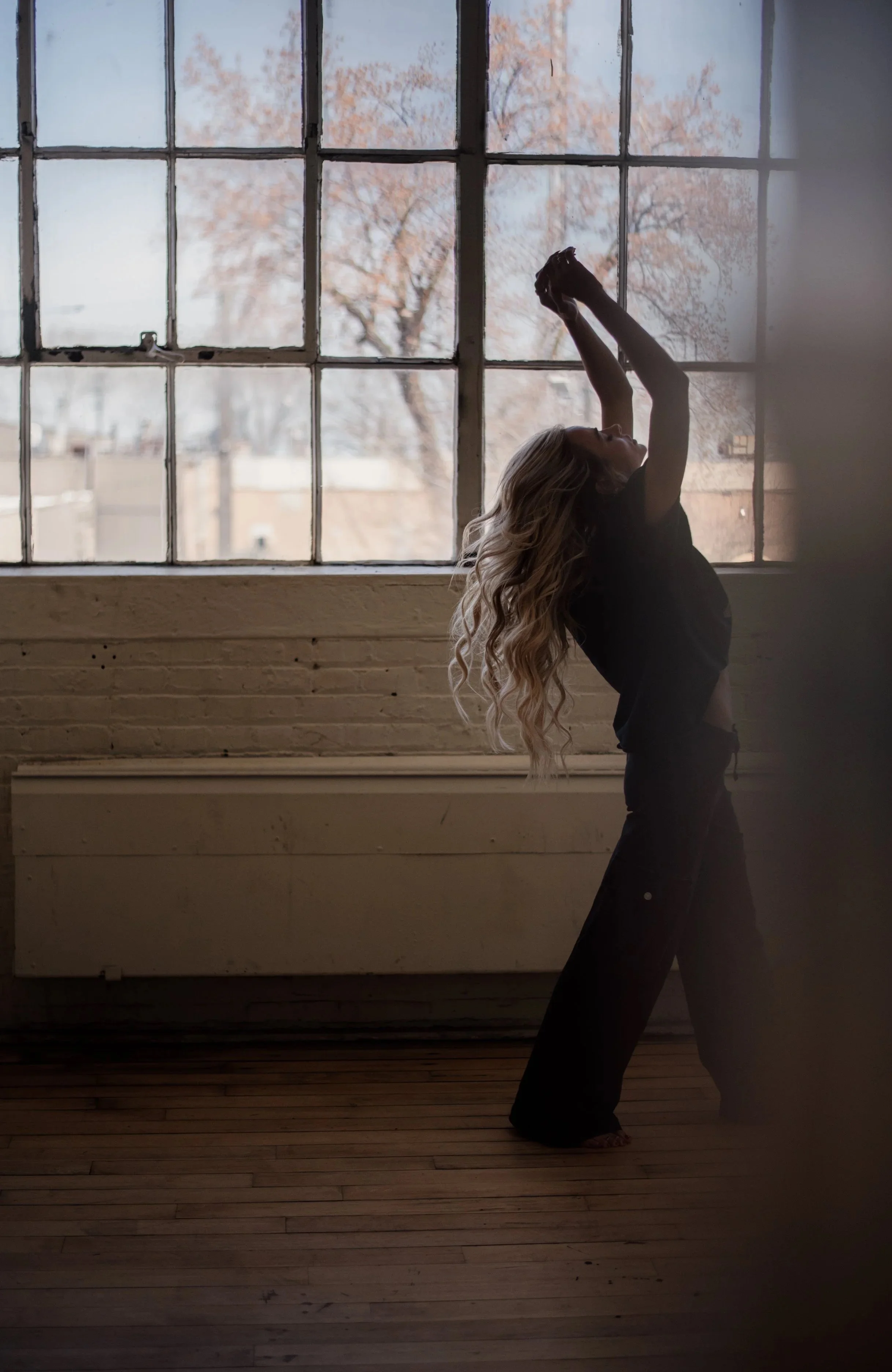 A woman with long wavy blonde hair stretching her arms upward while standing on a wooden floor in front of a large window with an outdoor view of trees and buildings.