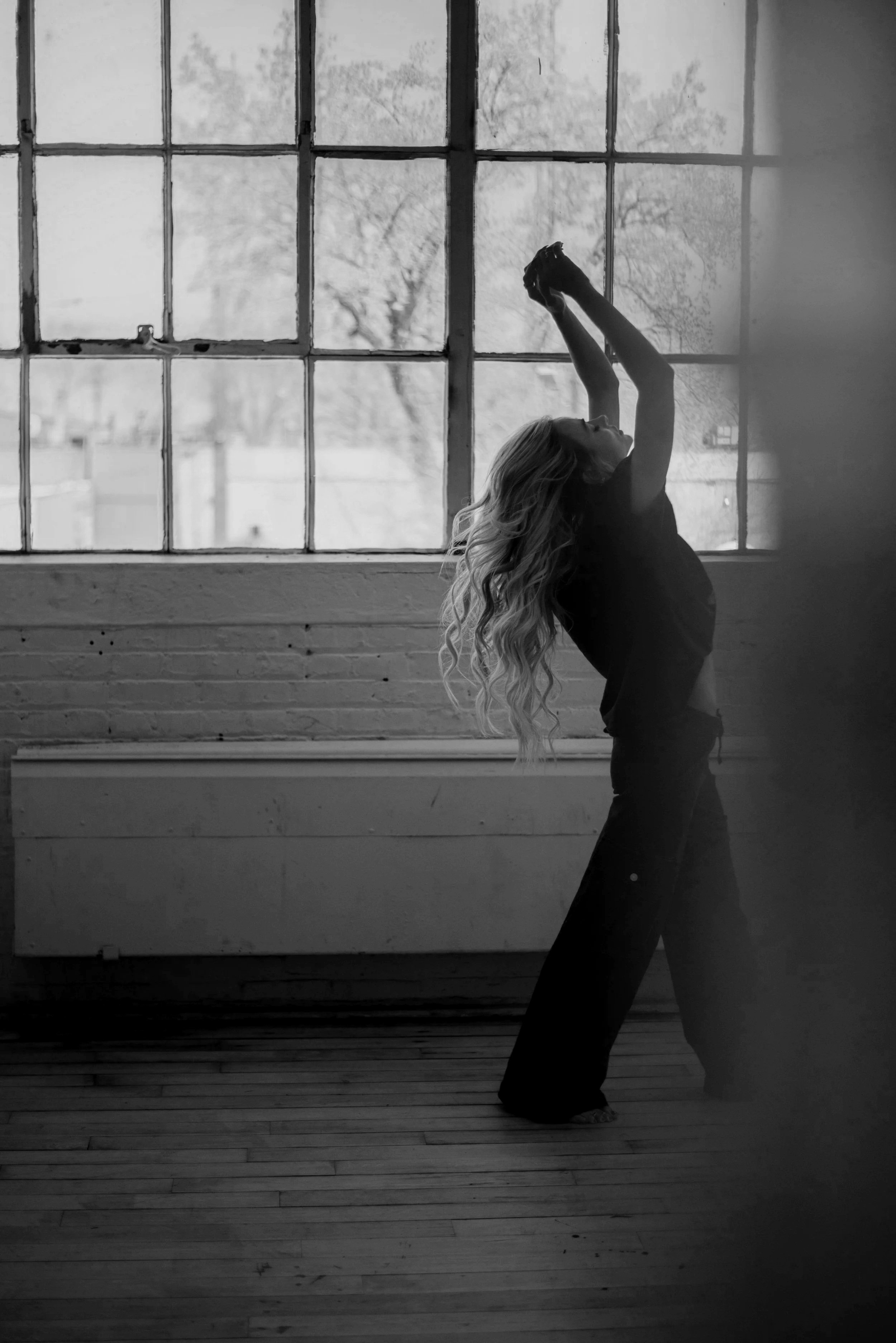 A woman with long wavy hair doing a yoga pose called the upward bow or wheel pose in front of a large window showing trees outside. She is standing on her feet with her hands reaching over her head, back arched, and head tilted back.