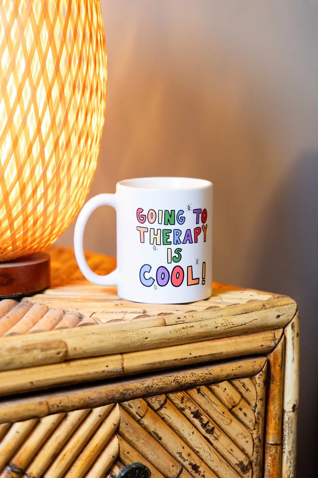 A white coffee mug with colorful text that says 'Going to therapy is cool!' and a lamp with a woven, orange shade on a wooden surface.