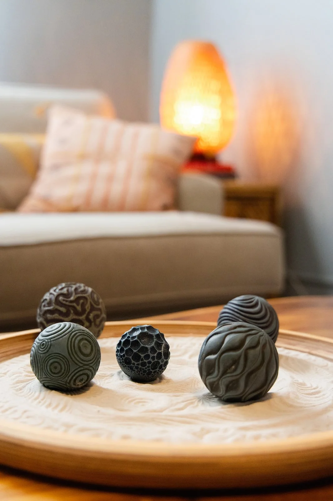 Five decorative black and gray marbles with various patterns on a wooden tray, with a blurred background of a beige sofa, plaid pillow, wooden side table, and a warm orange lamp.